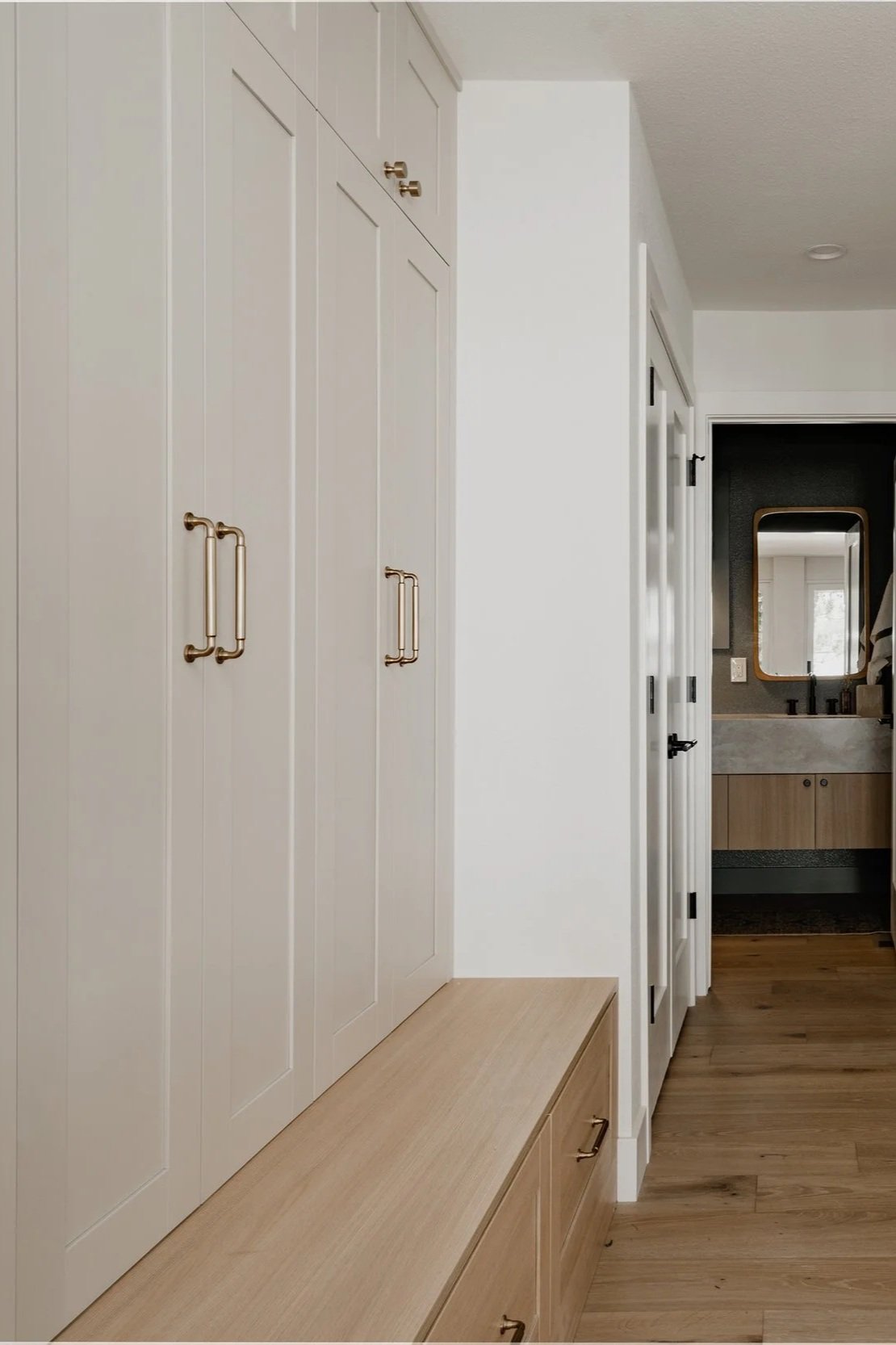 A hallway with built-in white cabinets with brass handles, a light wooden bench with drawers, and a view into a bathroom with a mirror and vanity sink.