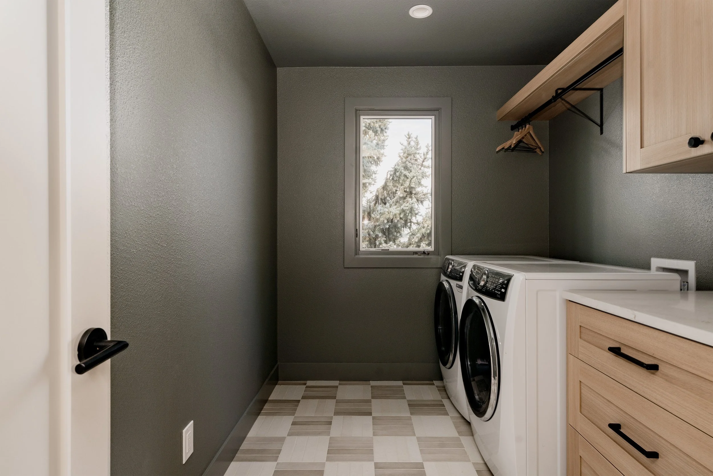 A laundry room with a window showing snow-covered trees outside, a washer and dryer, and wooden cabinets.