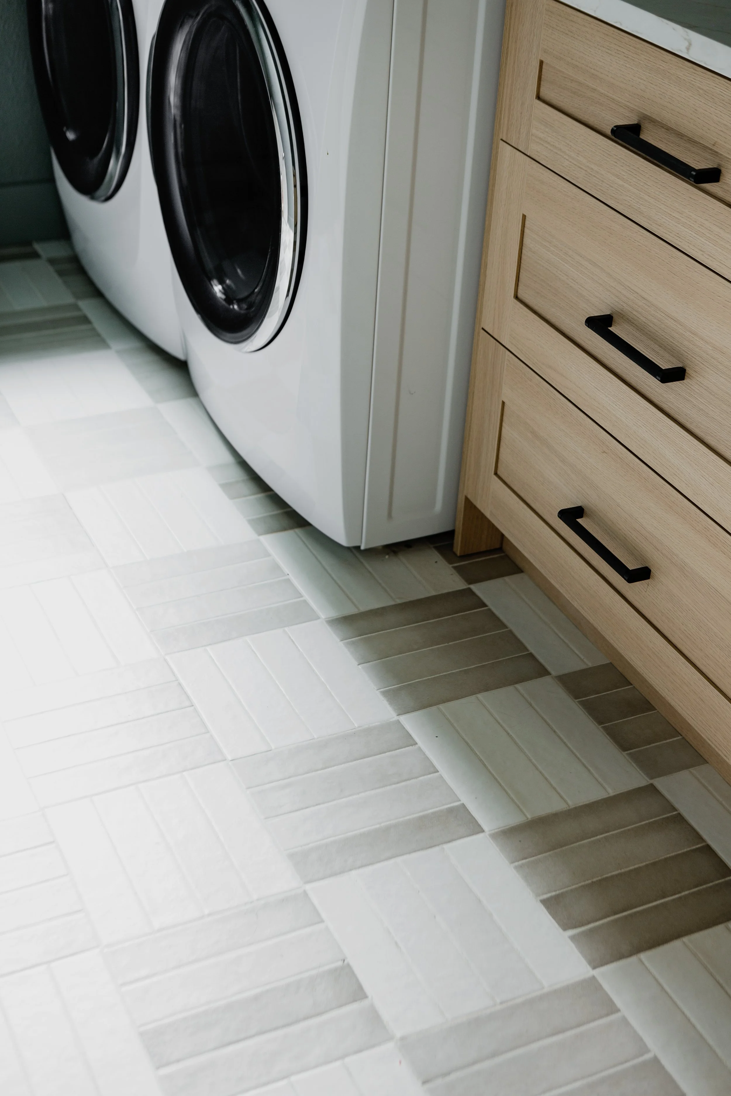 A laundry room with two front-loading washing machines, a light wood cabinet with three drawers, and tiled flooring in shades of light gray and beige.