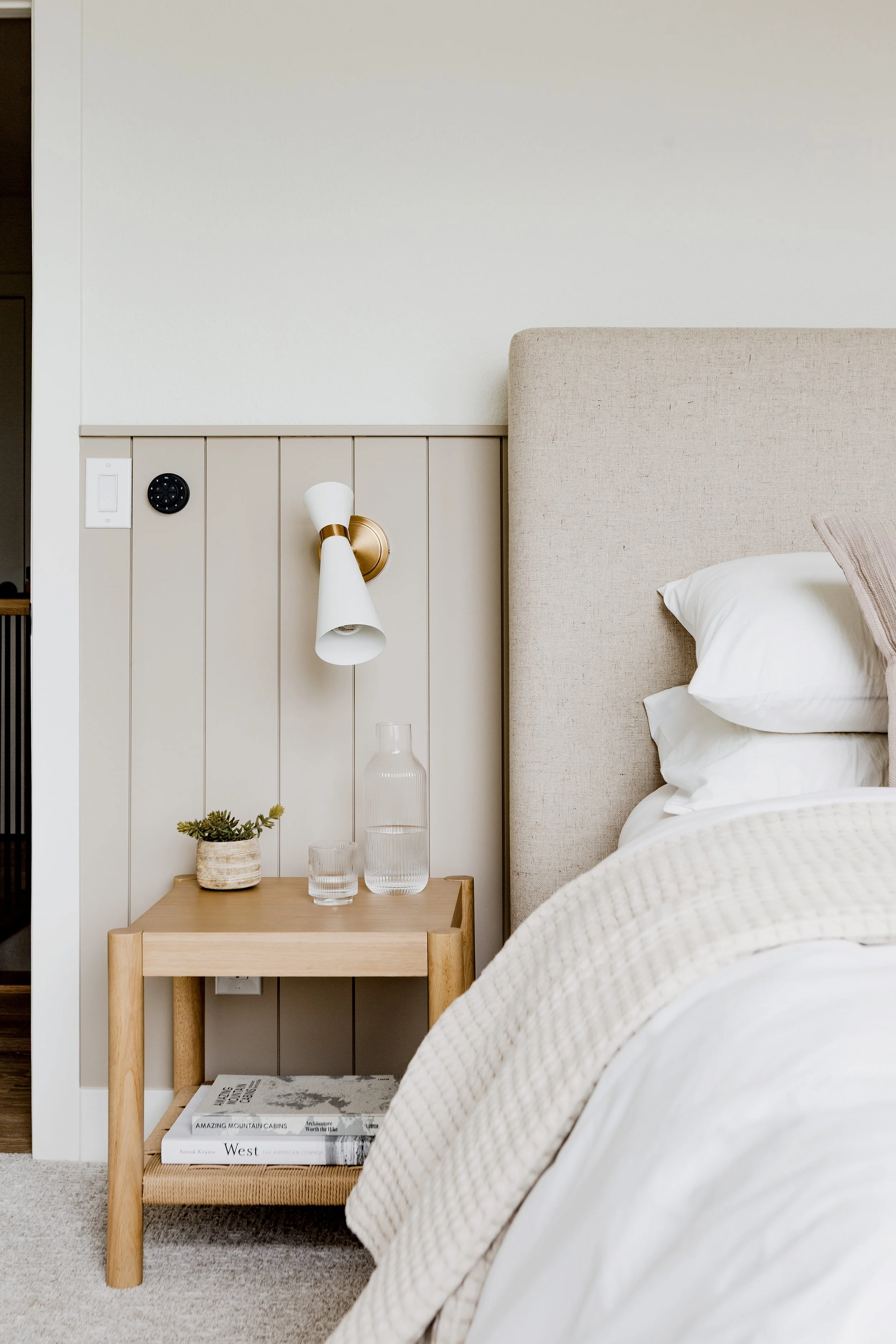 A bedroom with a beige upholstered headboard, white bedding, and a small wooden bedside table with glasses, a potted plant, and magazines. A wall-mounted lamp with a brass finish and white shade is above the table. The wall behind the bed is white with a wood panel section.