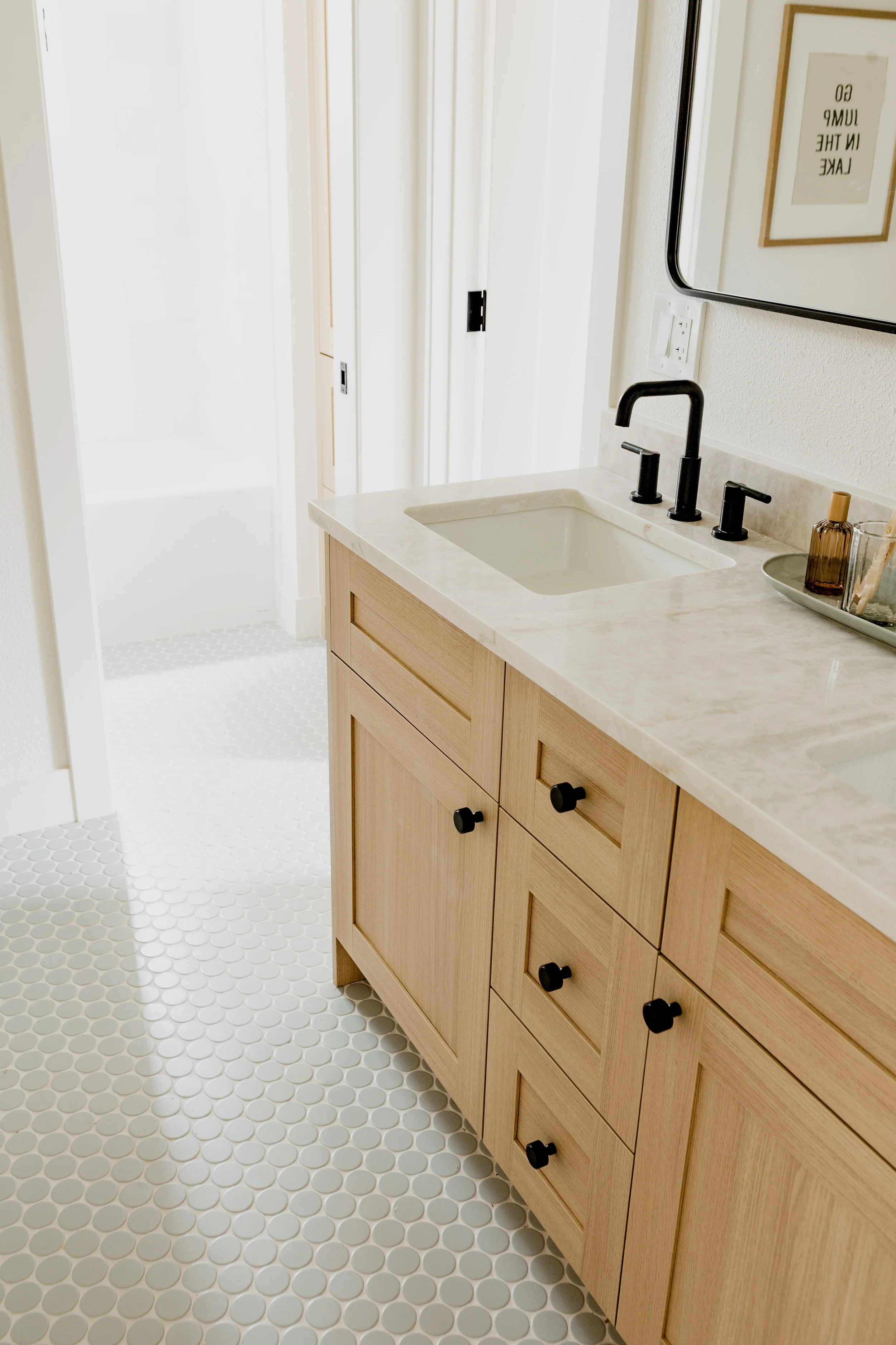 Bathroom with a light wood vanity with black knobs, a white countertop, and black faucet, mirror, framed art, and soap dispenser, white wall with a light switch, white hexagon floor tiles, and a doorway leading to a shower area.