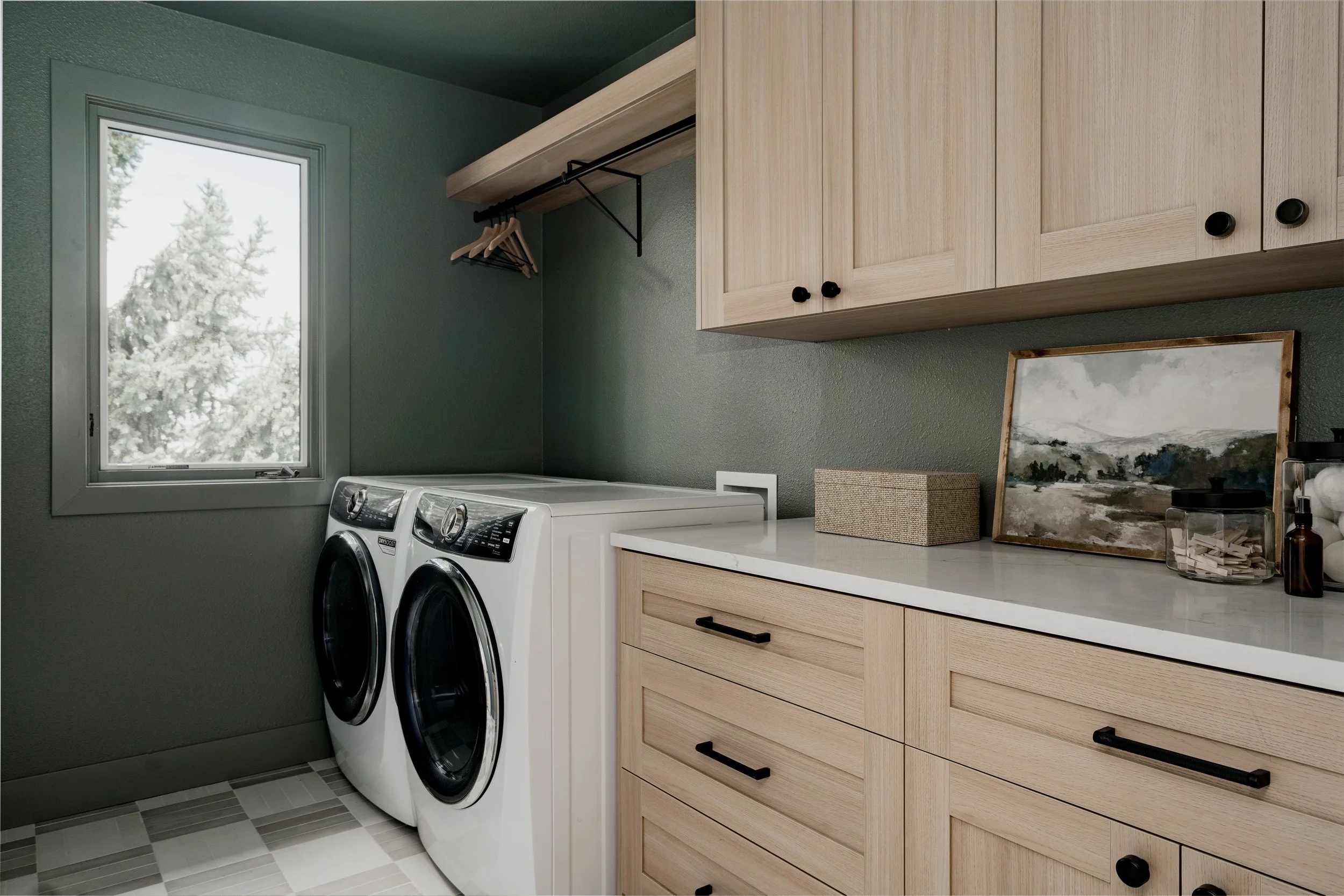 A laundry room with a window showing snow-covered trees outside. There are two front-loading washing machines side by side under a wooden shelf with hangers. To the right, wooden cabinets with black knobs are mounted on the wall, and a countertop below holds a framed picture, glass jars, and a small box.