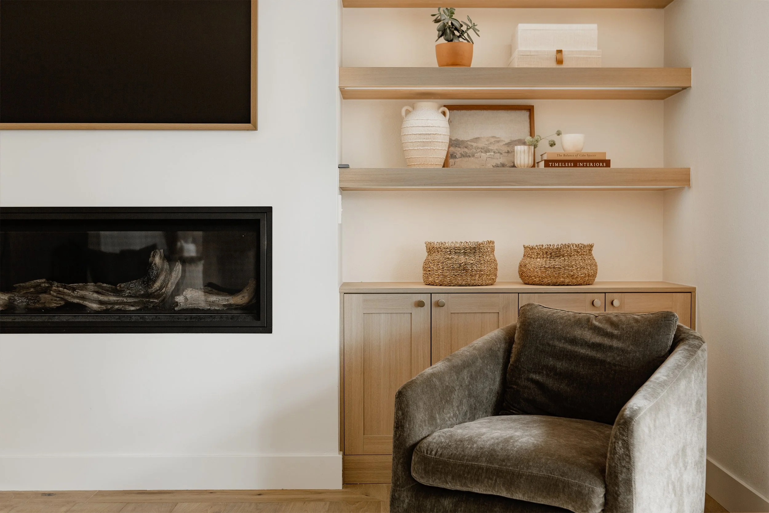 A living room with a black-framed fireplace on a white wall, wooden floating shelves with decorative items, and a plush gray armchair.