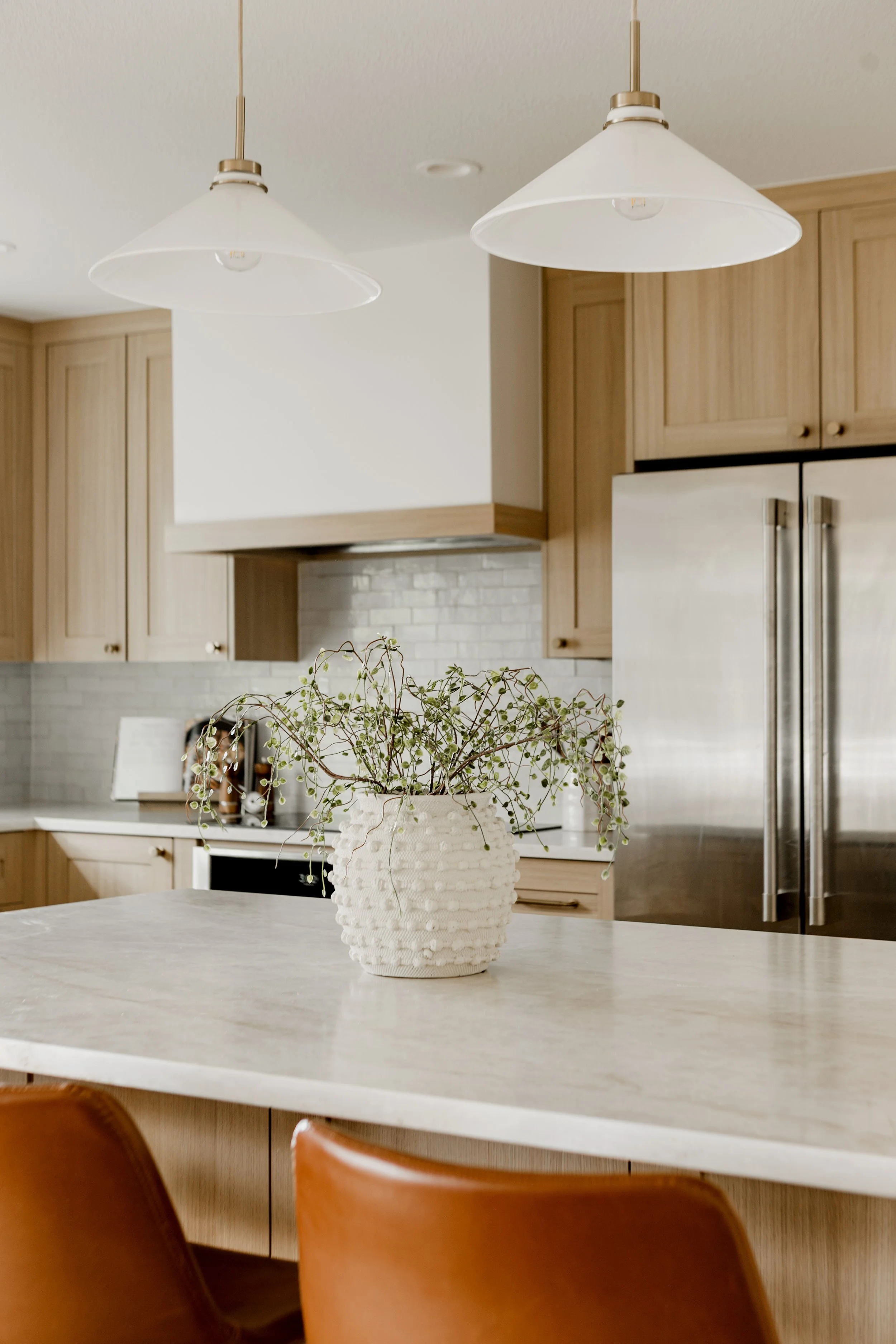 A kitchen interior with a white vase containing a green plant on a light-colored countertop, wooden cabinets, stainless steel refrigerator, and two white pendant lights hanging from the ceiling.