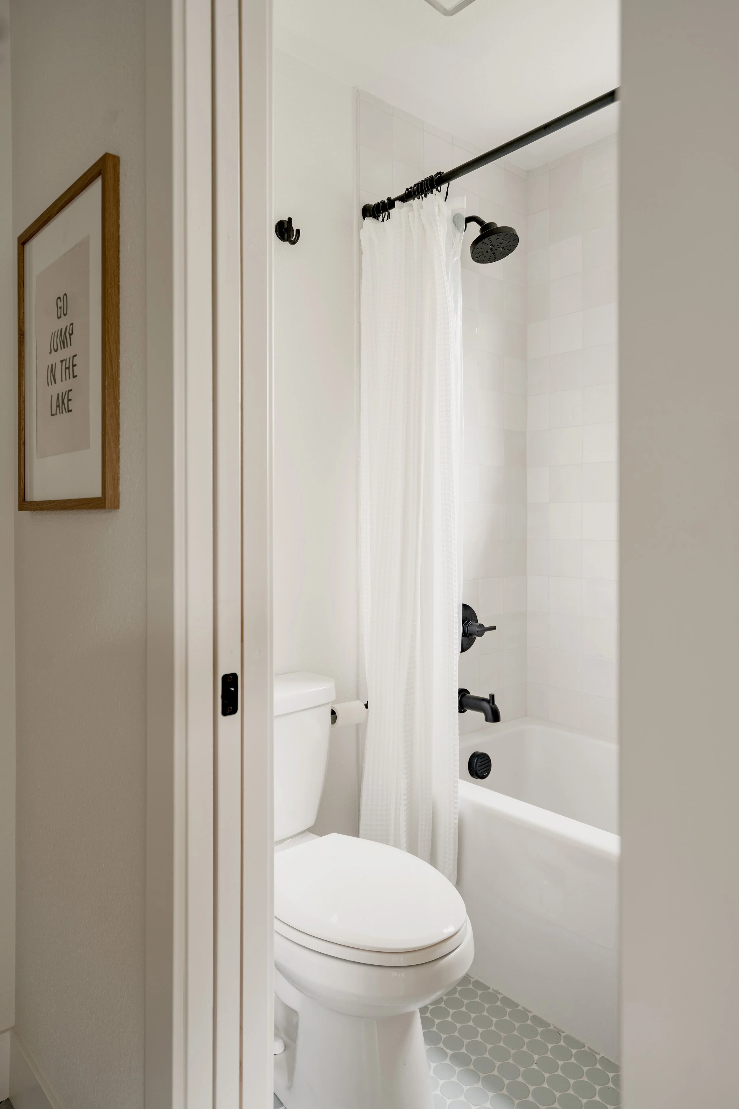Small bathroom with a white toilet, a tub with a shower, white shower curtain, black shower head and fixtures, white tiled walls, and a gray hexagonal tile floor. Part of a framed art print on the wall reads 'Go Jump in the Lake.'