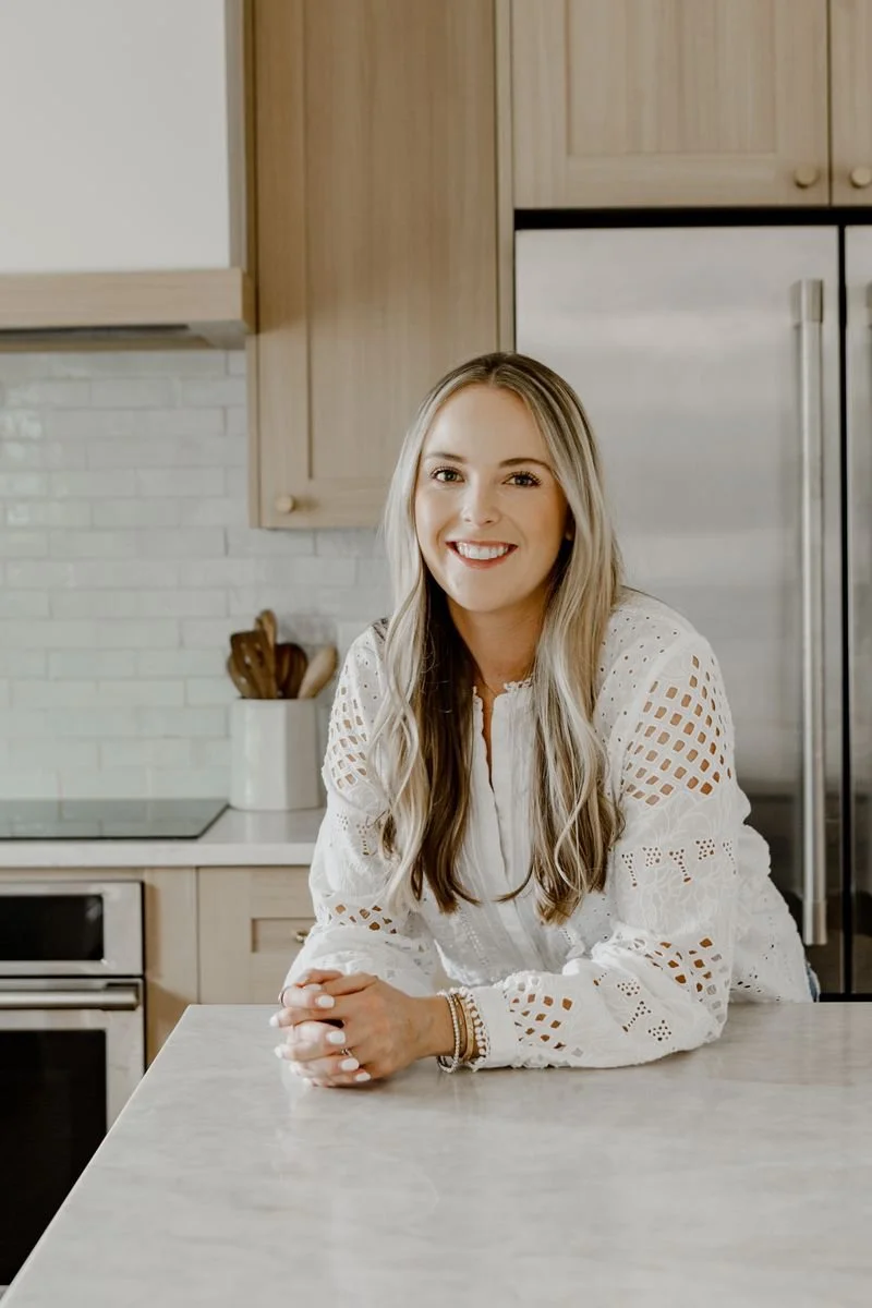 A smiling woman with long blonde hair, wearing a white eyelet blouse, sitting at a kitchen counter in a modern kitchen with wooden cabinets and a stainless steel refrigerator.