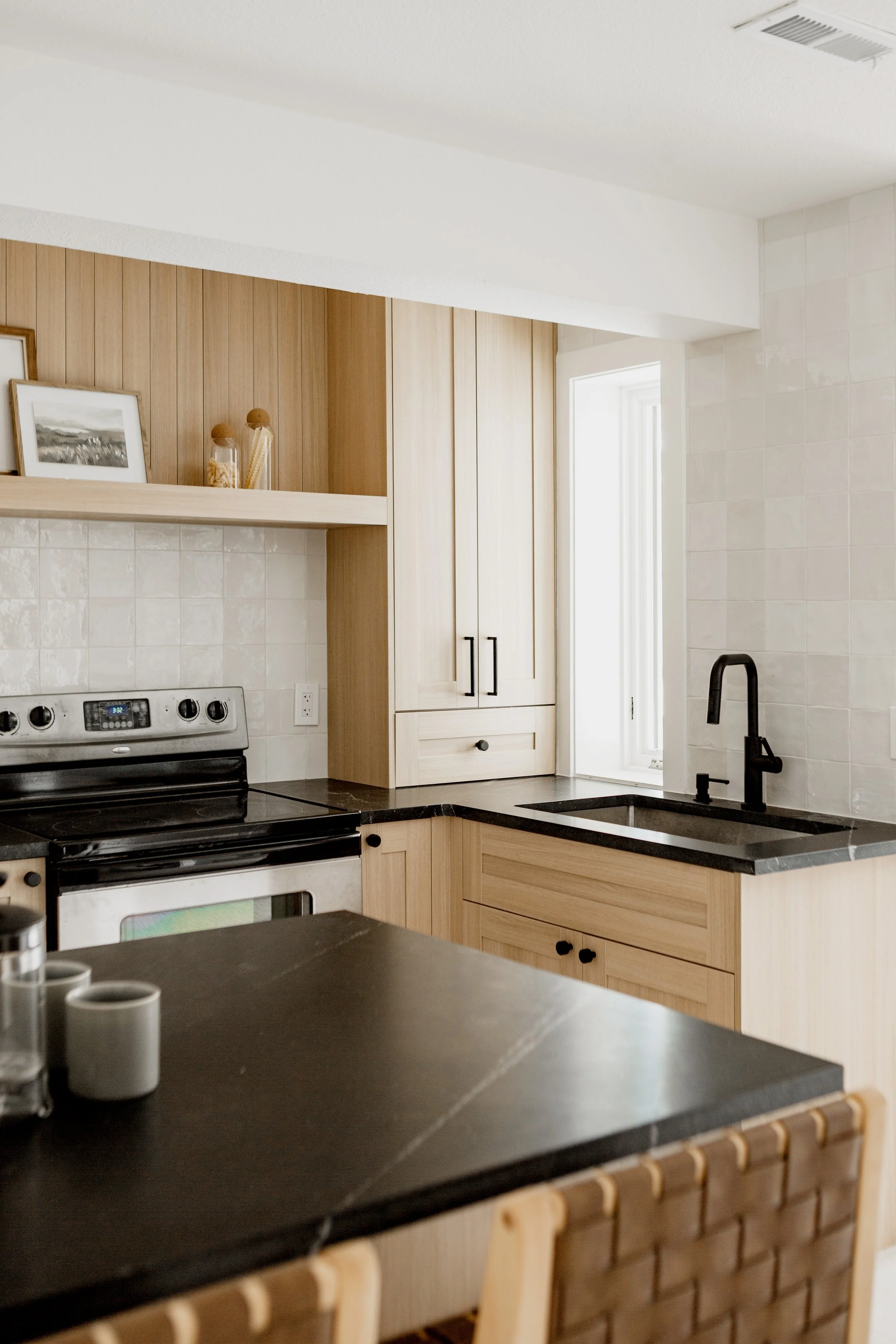 Modern kitchen with light wood cabinets, black countertops, a black faucet, and a white tiled wall. Some framed artwork and jars are displayed on an open shelf above the stove. There is a window allowing natural light.