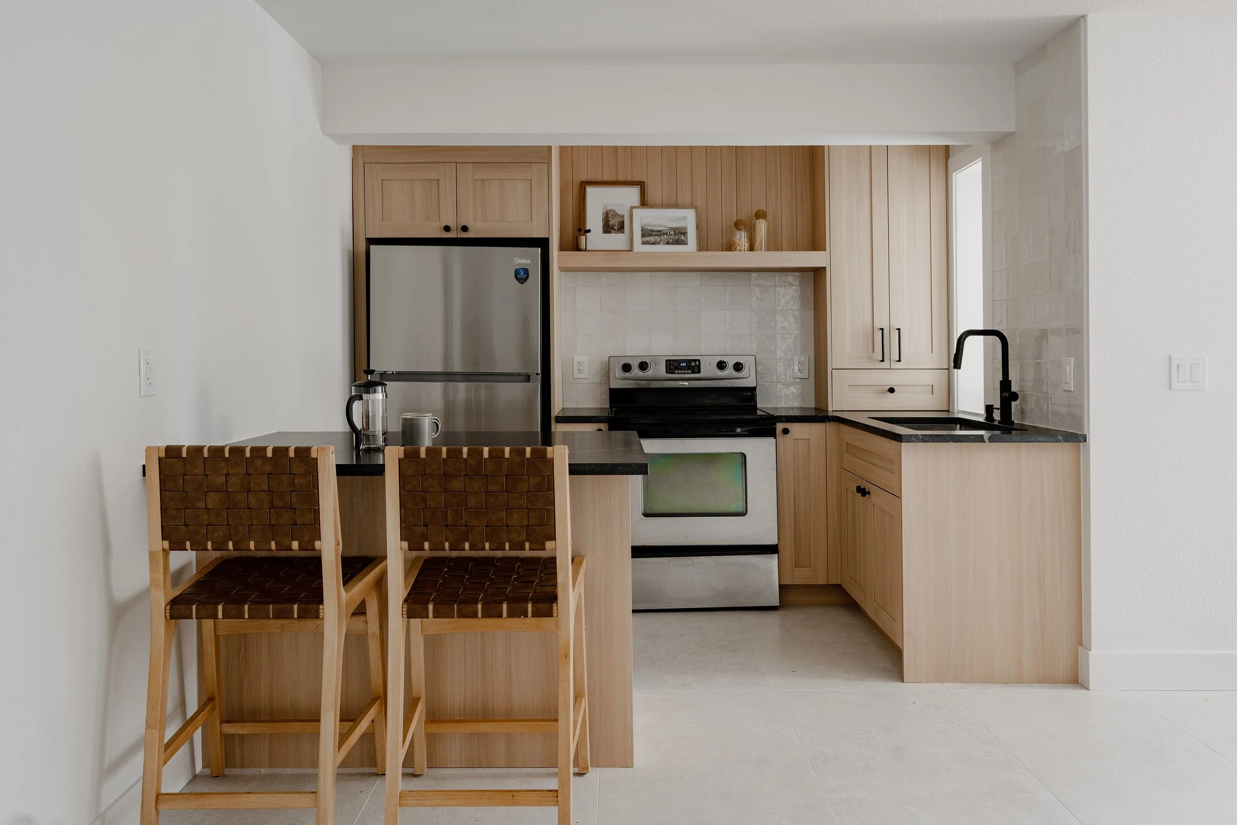A modern kitchen with wooden cabinets, black countertop, stainless steel refrigerator, black sink faucet, white walls, and delicate framed artwork above the stove. Two wooden chairs with woven seats are partially visible in the foreground.