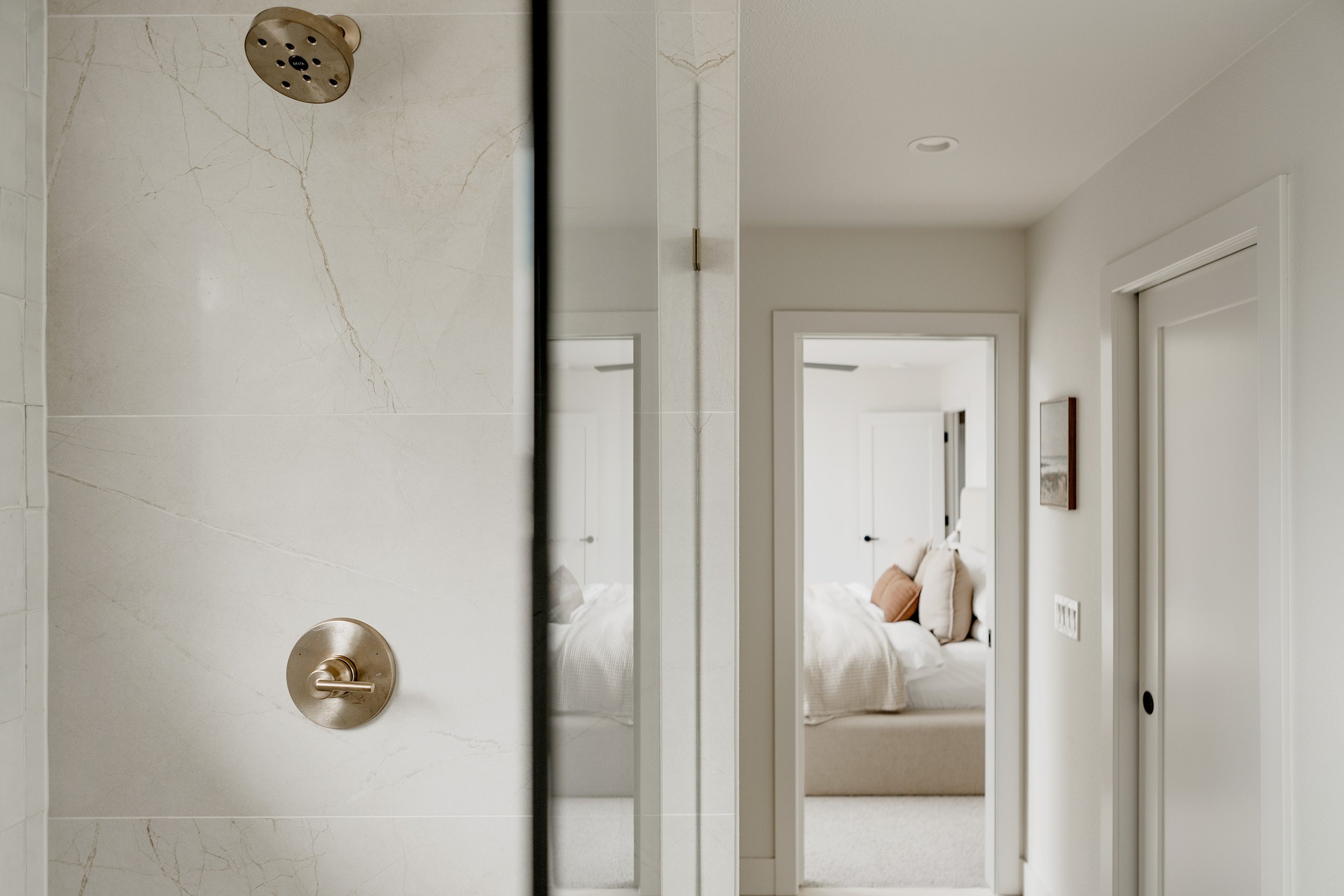 Bathroom shower with beige marble wall and gold showerhead, mirror reflecting bedroom with bed and wall art.