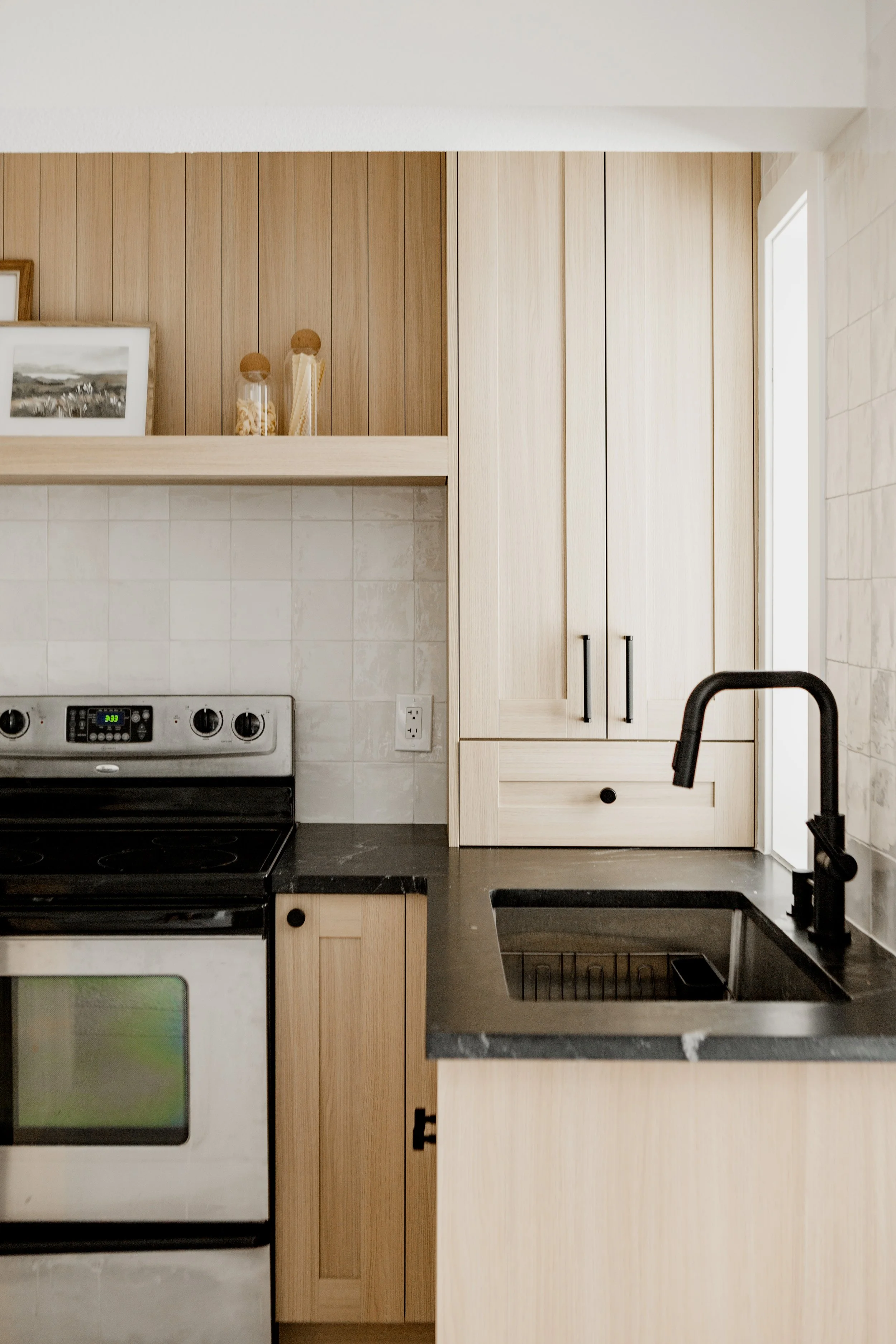 A kitchen with light wood cabinets, a black granite countertop, a stainless steel sink, a black faucet, a stove with control knobs, an electric outlet, a framed photo on a shelf, and decorative jars.
