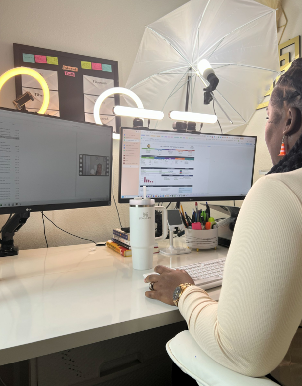 A woman working at a desk with two computer monitors, a white water bottle, a notebook, and assorted pens and pencils, with a white photography umbrella and lights in the background.