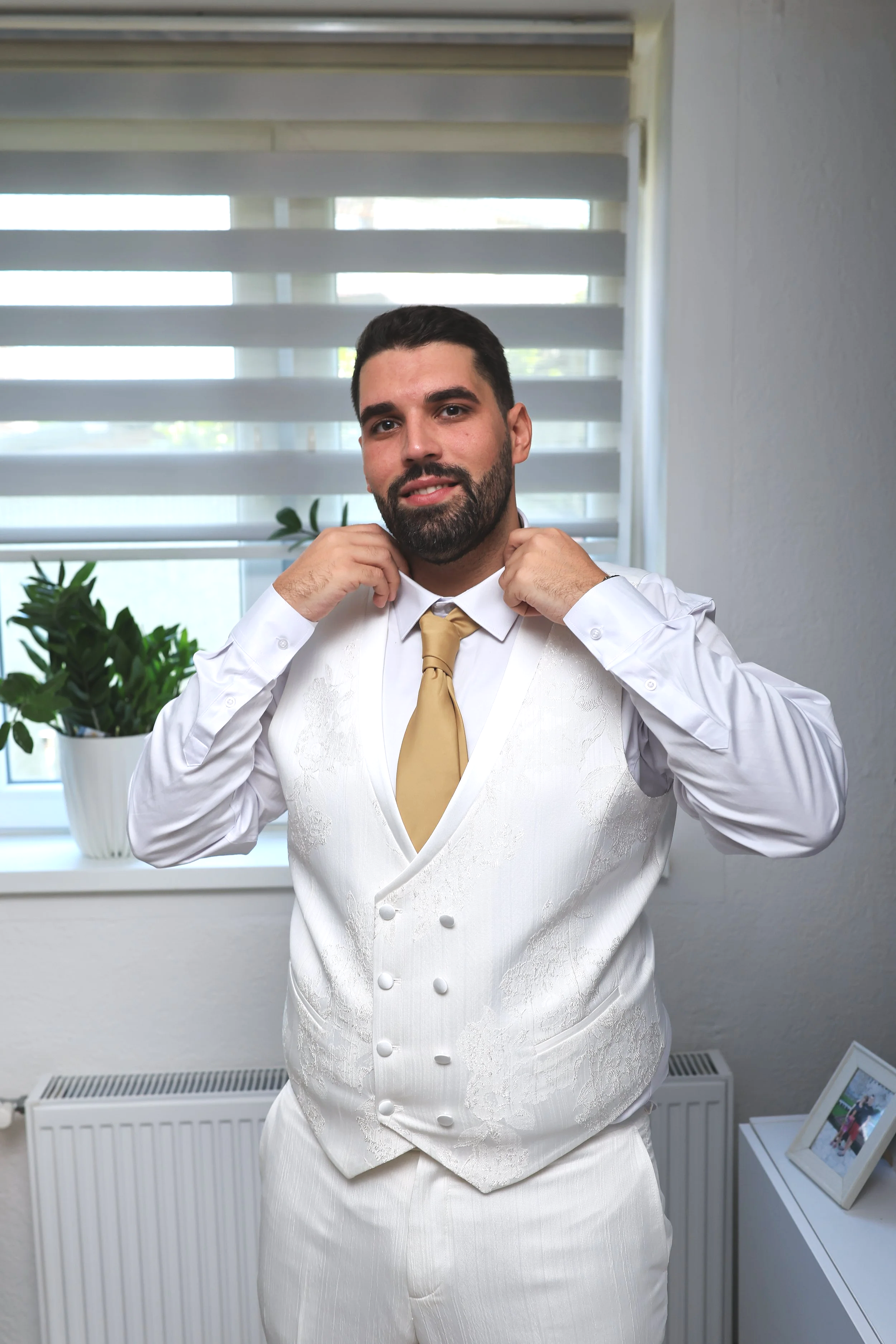 A man with dark hair and a beard adjusting his white vest and tie in front of a window with horizontal blinds, with a potted plant and framed photos on the windowsill in the background.