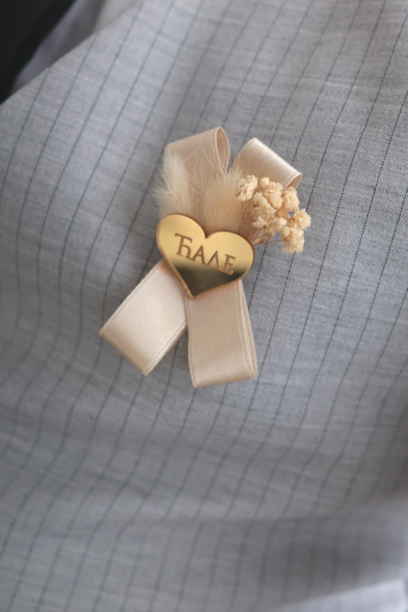 A close-up view of a boutonniere on a lapel, consisting of a cream-colored flower, beige ribbon, white feather, and a gold heart with the word 'ΤΑΛΕ' written on it in Greek.