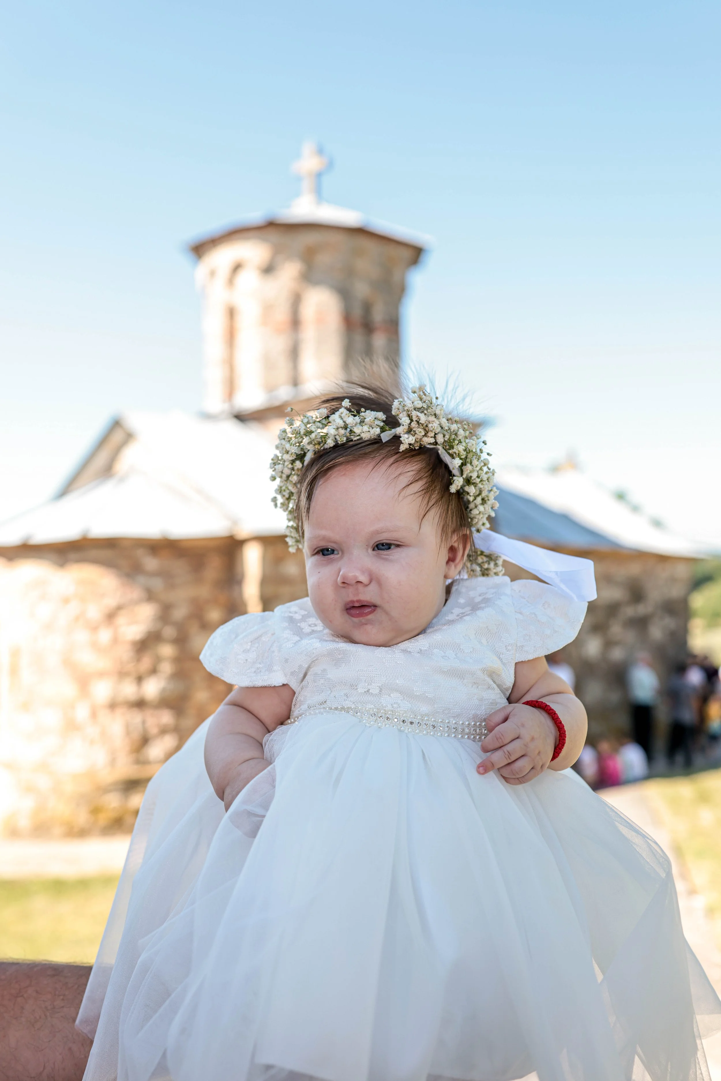A young girl wearing a white dress and a flower crown, being held up outdoors near a church with a stone tower, blue sky, and a crowd of people in the background.