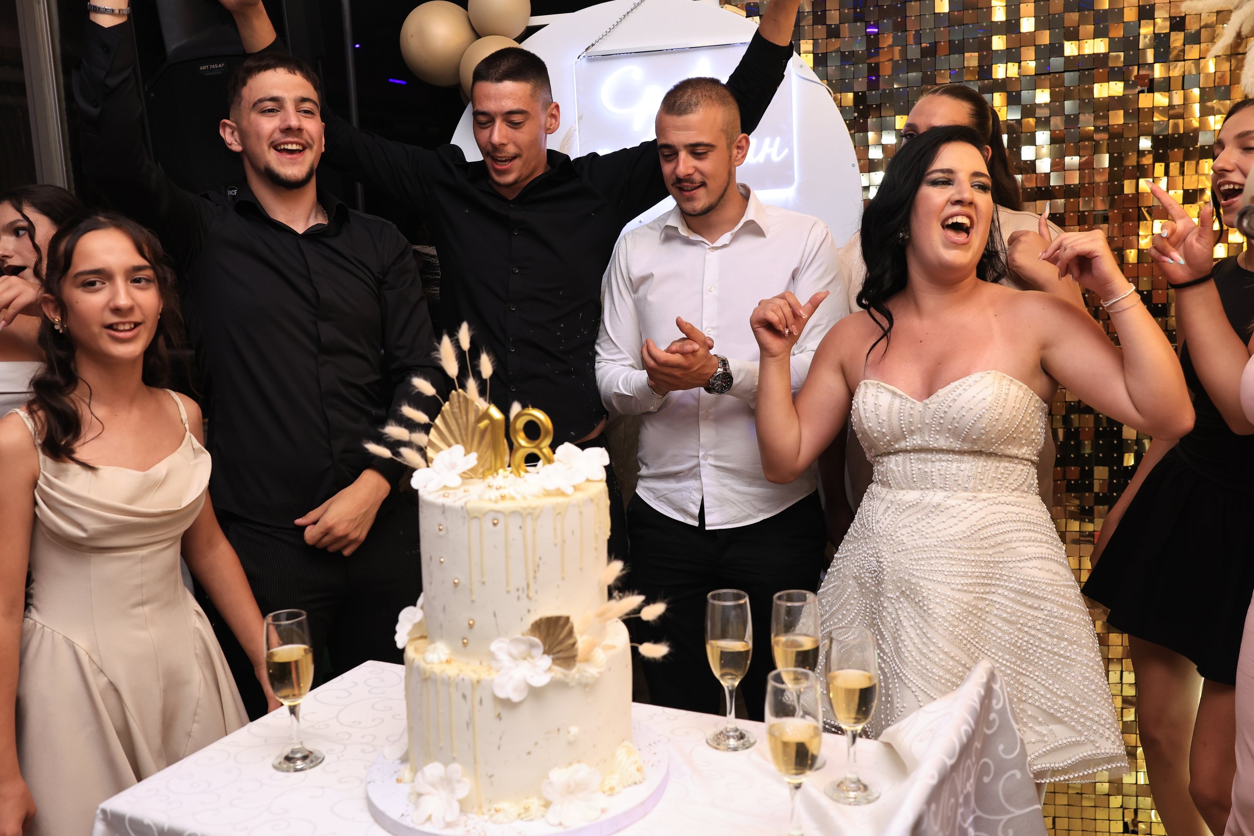 A group of people celebrating a special occasion, with a cake, champagne glasses, and a gold and black decor background.