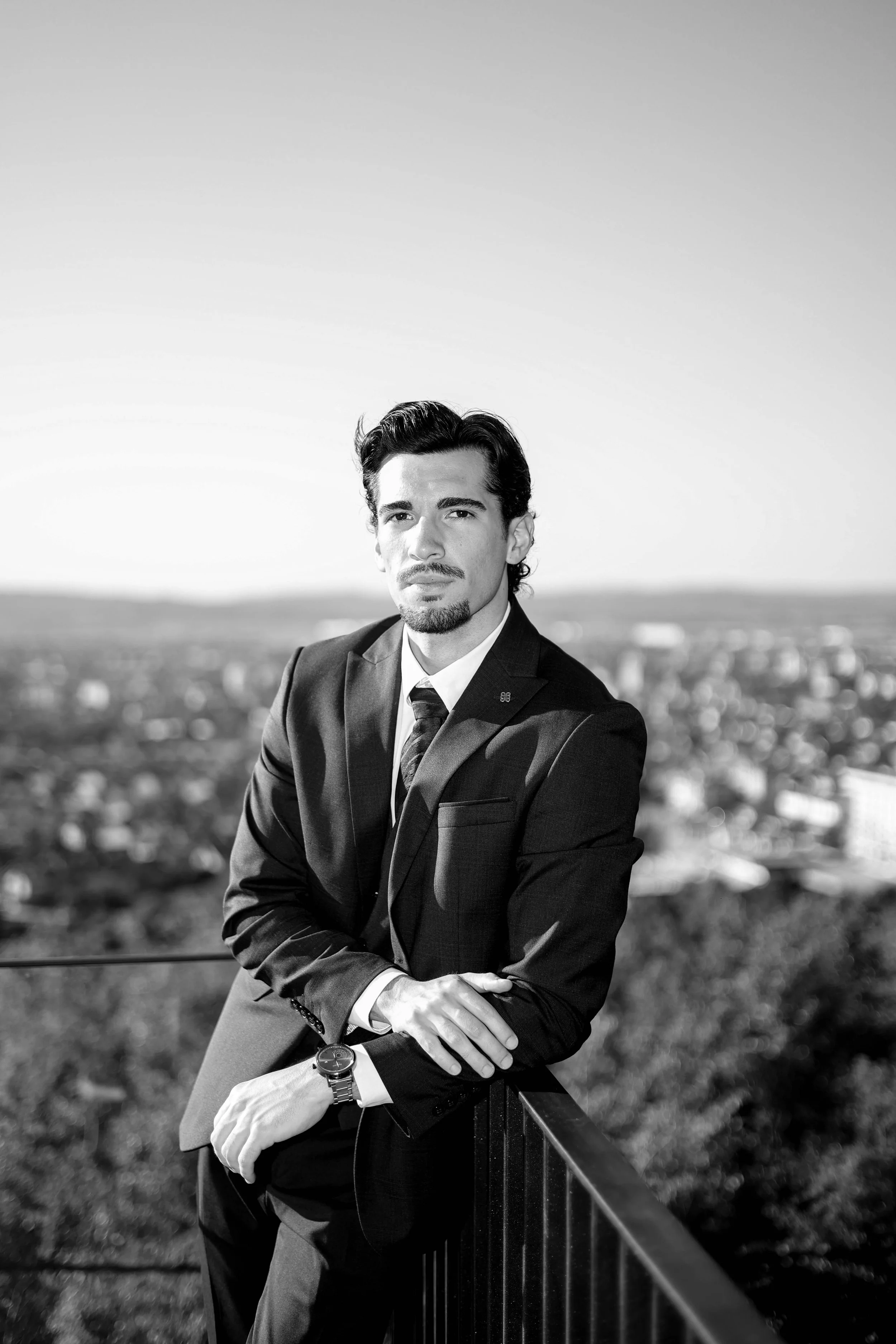 A man with dark hair and a mustache dressed in a suit, standing outdoors on a balcony with a cityscape in the background.