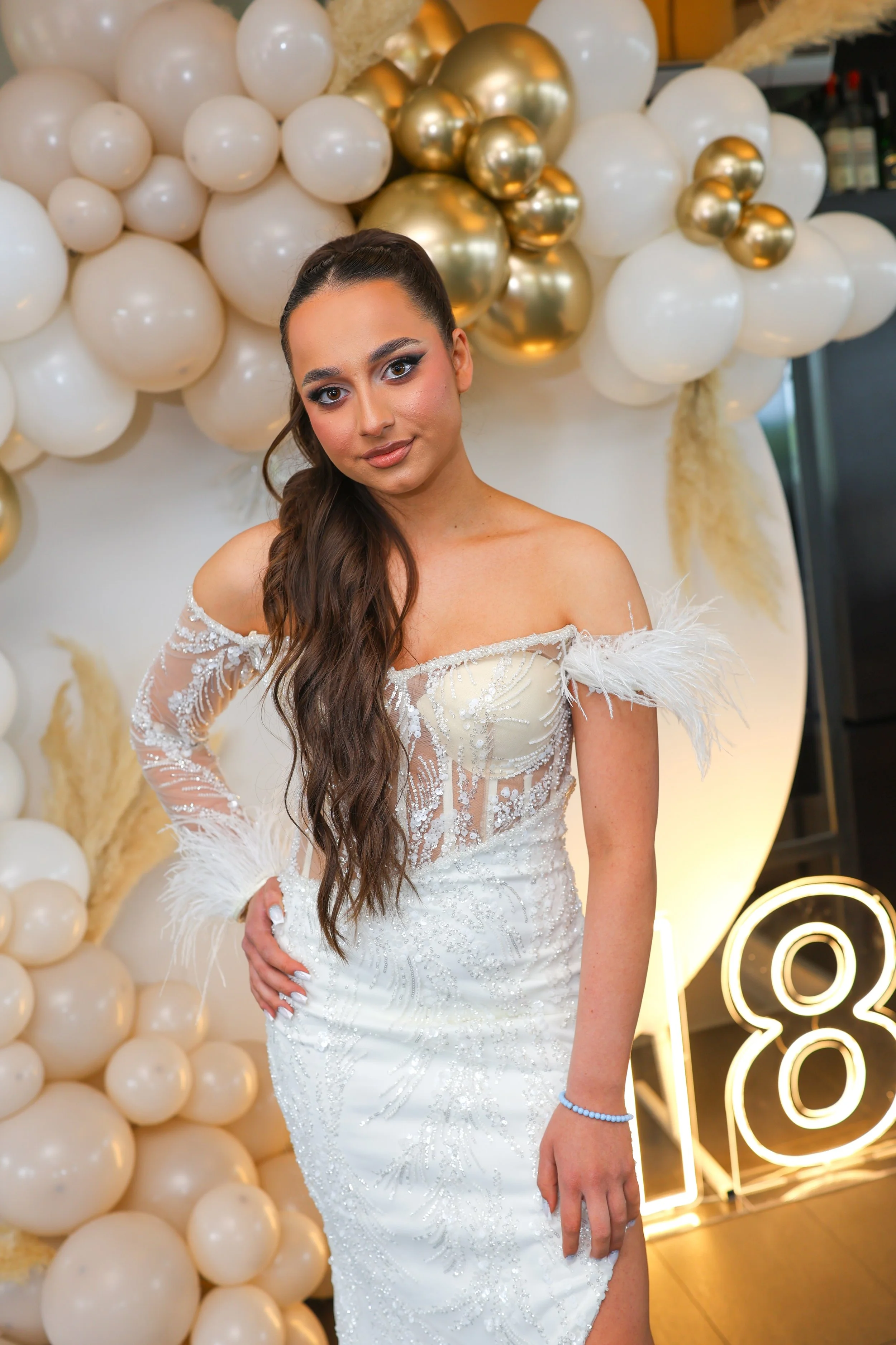 A young woman in a white wedding dress with feather accents on the sleeves, standing in front of a backdrop of white and gold balloons and a neon light display.