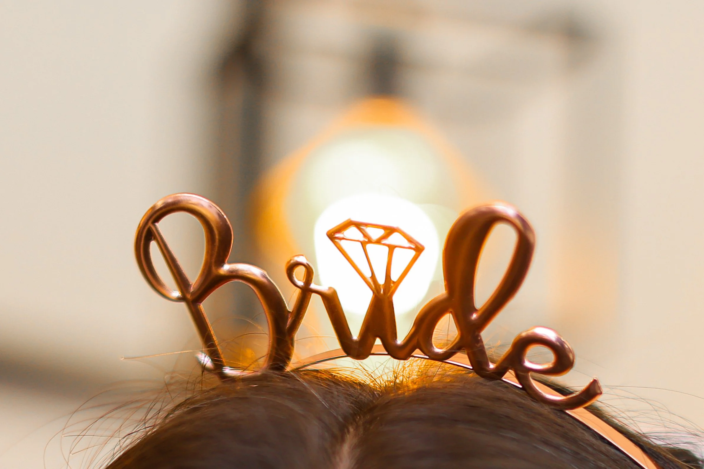 Close-up of a person's head with brown hair, wearing a metallic hair accessory resembling a crown with a diamond shape in the center, against a blurred background with warm lighting.