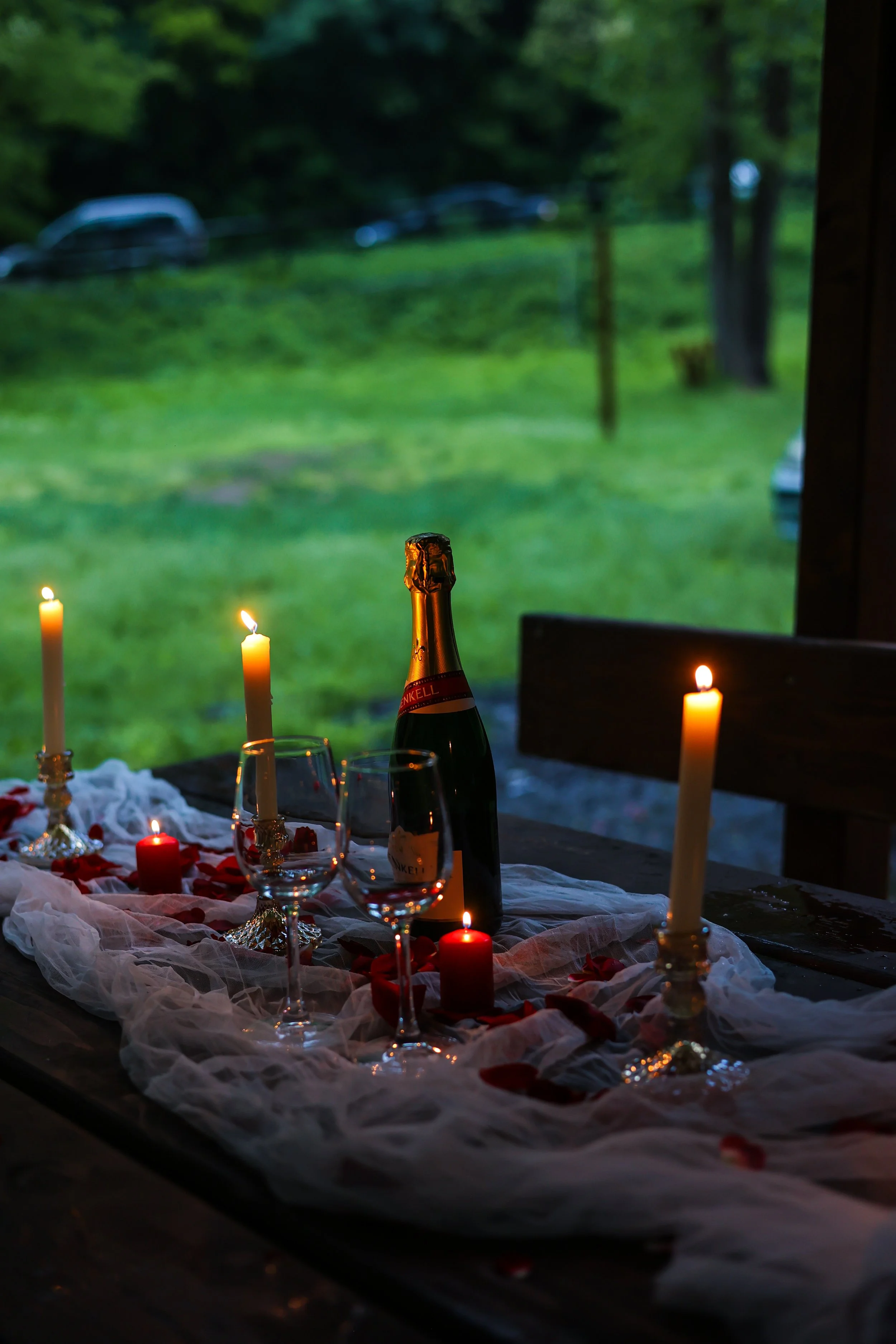 A table decorated with candles, rose petals, and a wine bottle, set for a romantic dinner, with a blurred green outdoor scene visible through a window in the background.