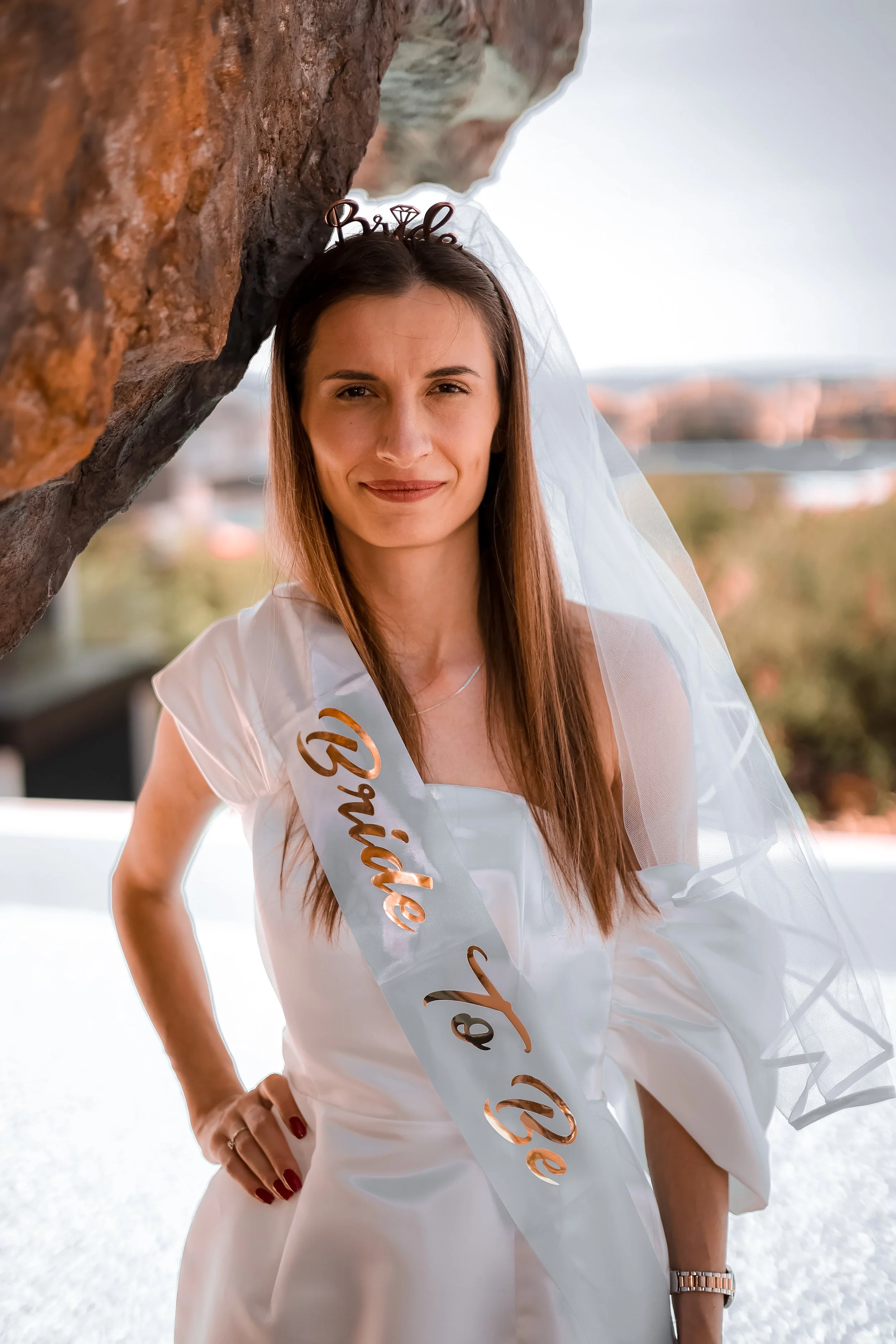 A woman wearing a white dress and a sash that says 'Bride to Be' with a veil and a headband that says 'Bride' is standing outdoors near a large rock formation. She has long brown hair, is smiling slightly, and has her left hand on her hip. The backgr