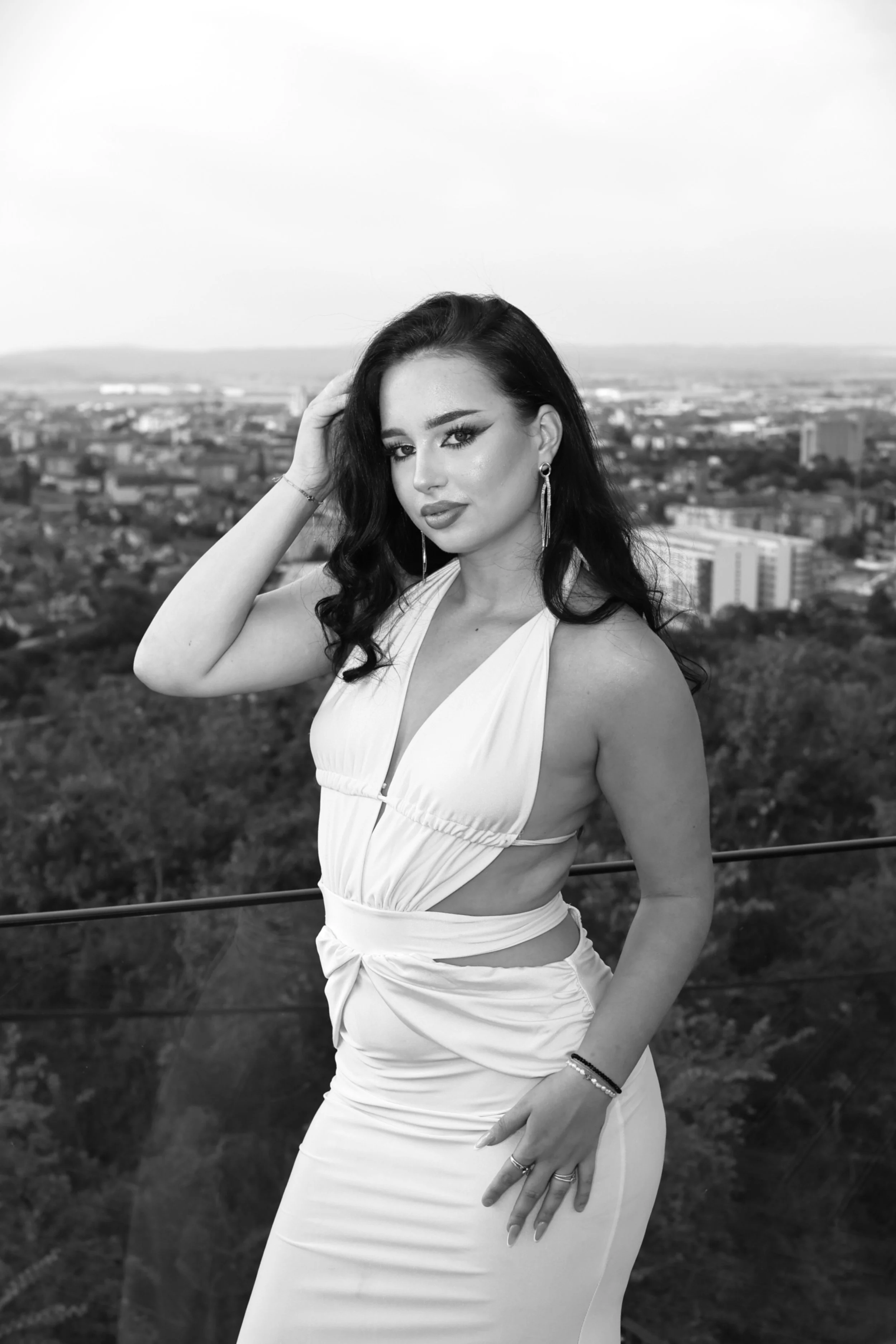 A woman with dark hair wearing a white sleeveless dress, standing outdoors on a cityscape background, looking at the camera, black and white photo.