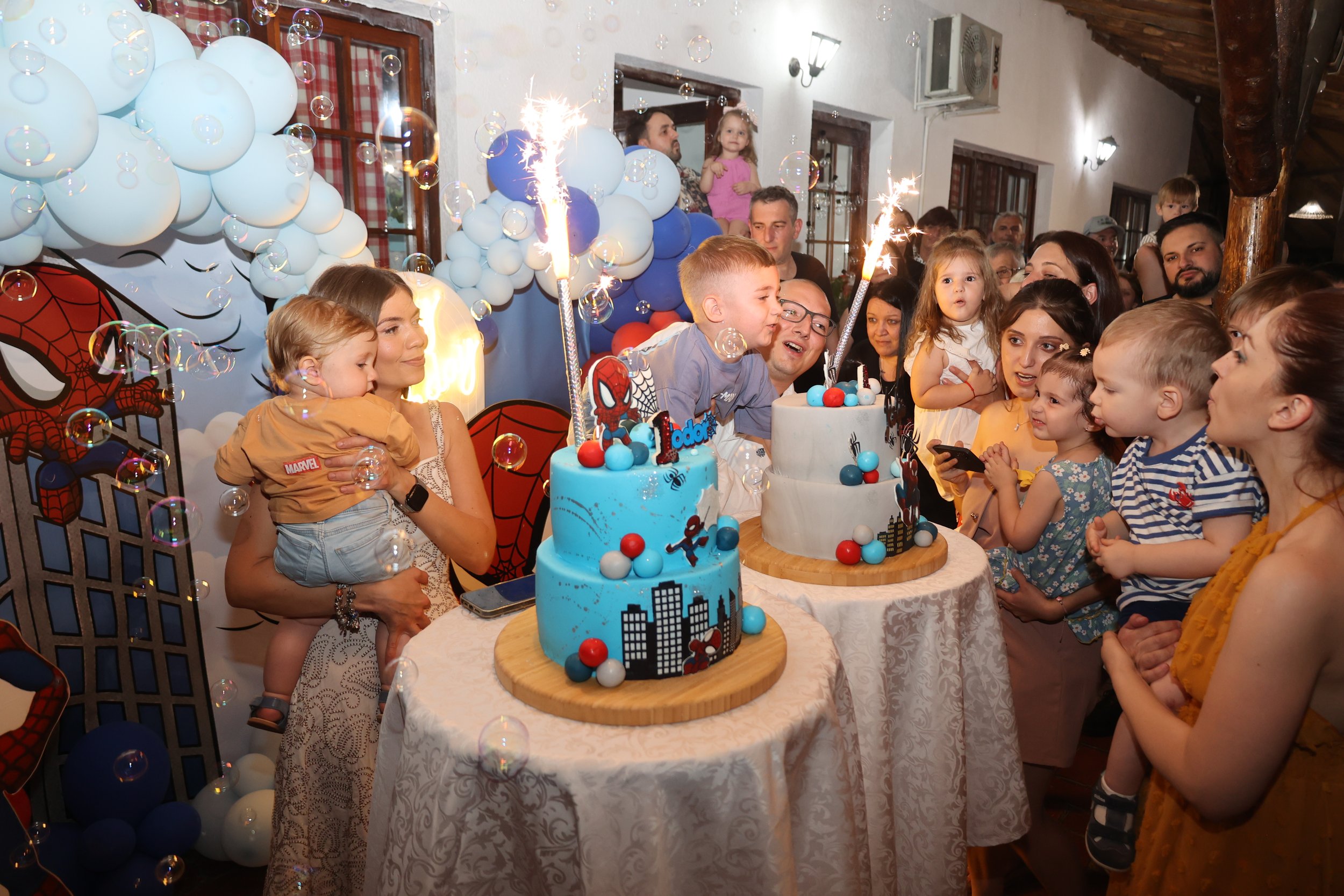 A children's birthday party with a superhero theme, featuring a decorated table with two colorful cakes, children and adults celebrating, balloons, bubbly decorations, and sparklers on the cakes.