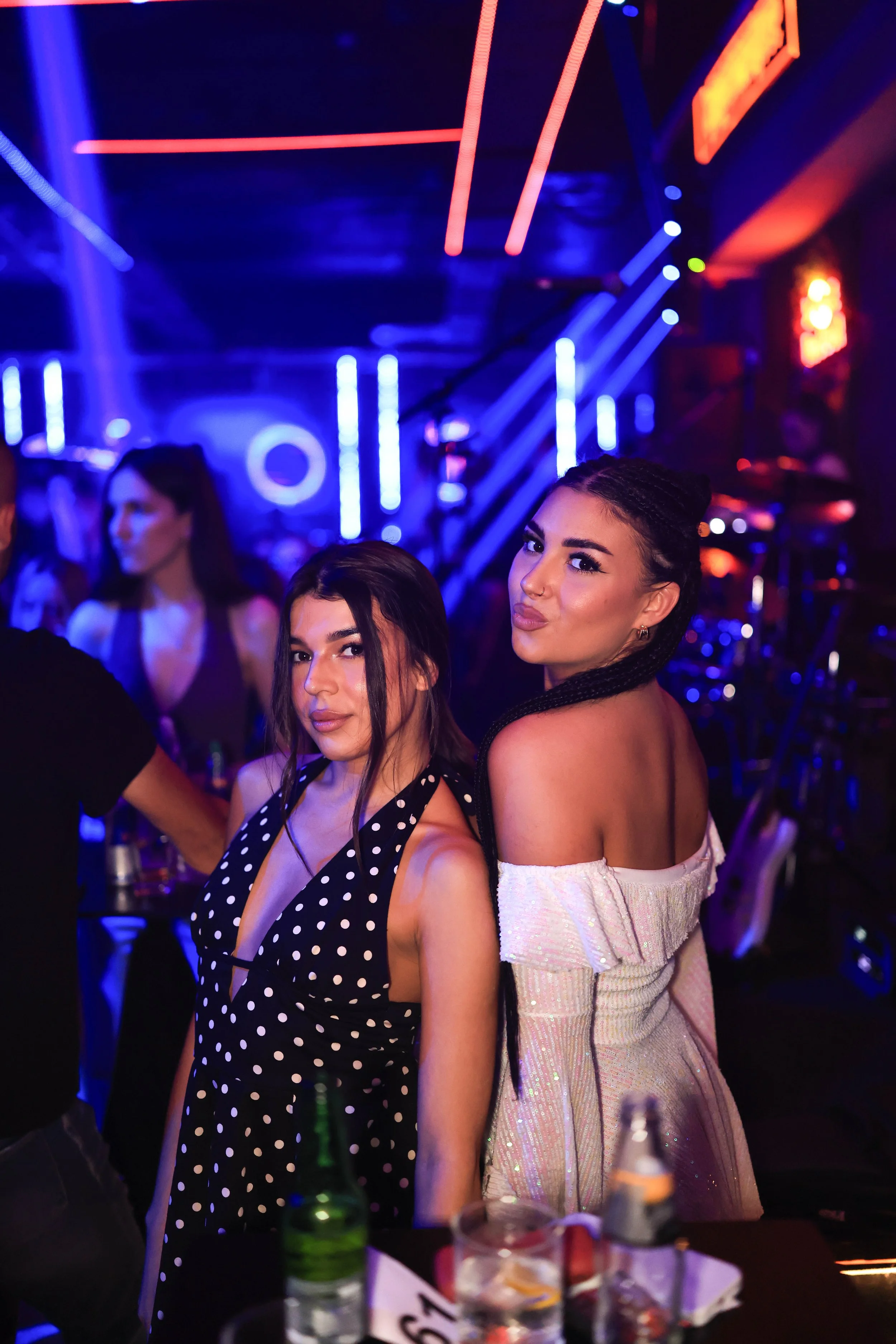 Two women, one with dark hair in braids and the other with long, dark hair, posing at a nightclub with colorful neon lights and other people in the background.