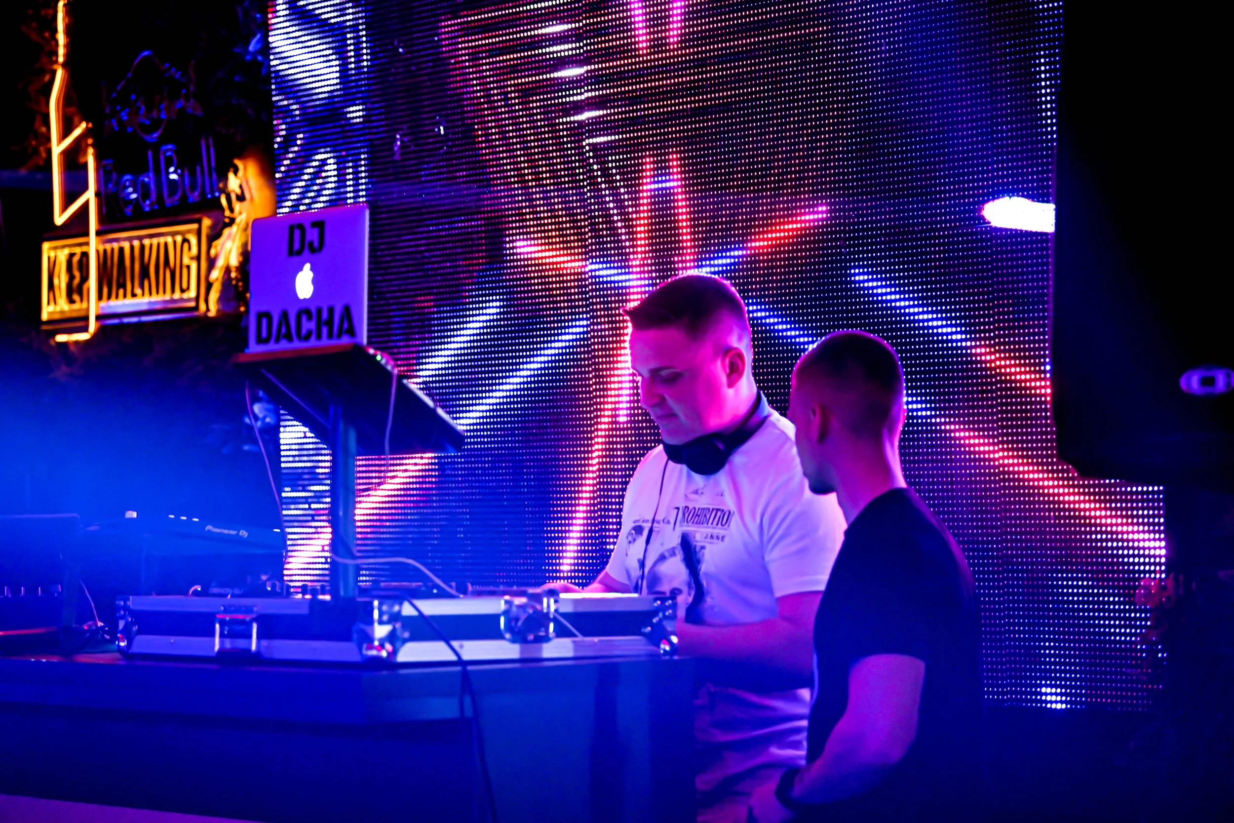 Two male DJs performing on a stage with bright, colorful LED lights and digital screens in the background at a nightclub or music event.