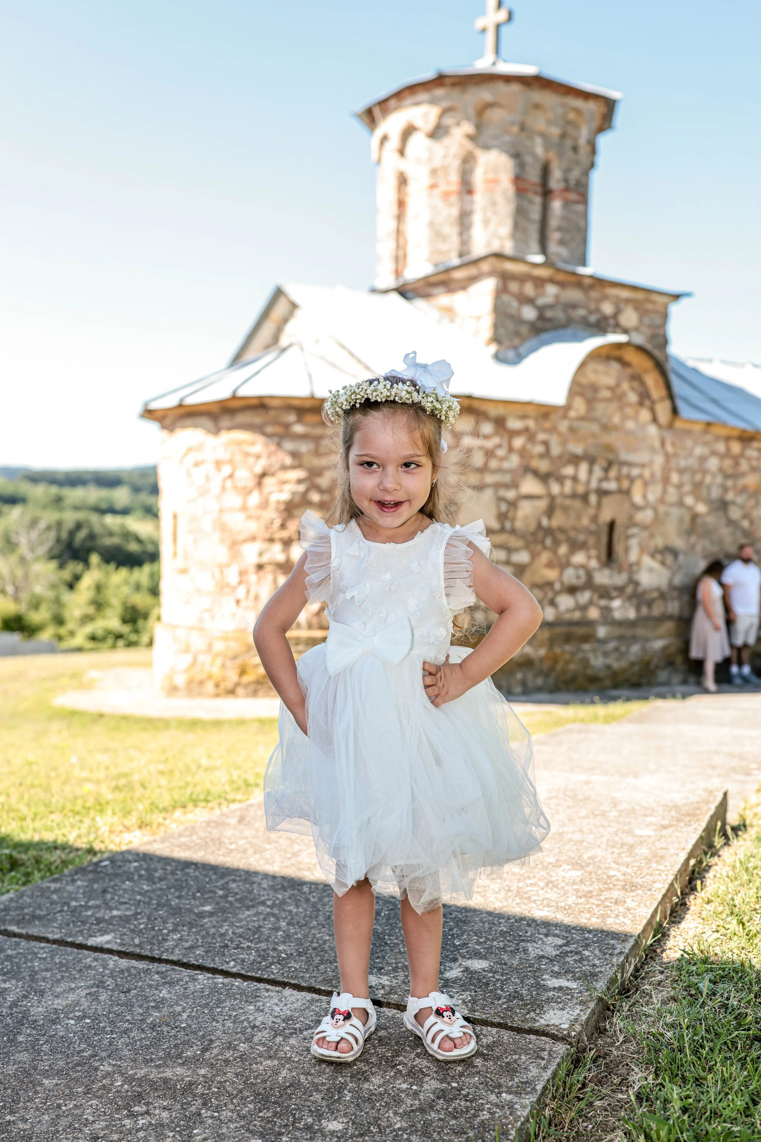 Young girl in a white dress with ruffled sleeves and a floral headband standing outdoors in front of a stone church with a cross on top, during a sunny day.
