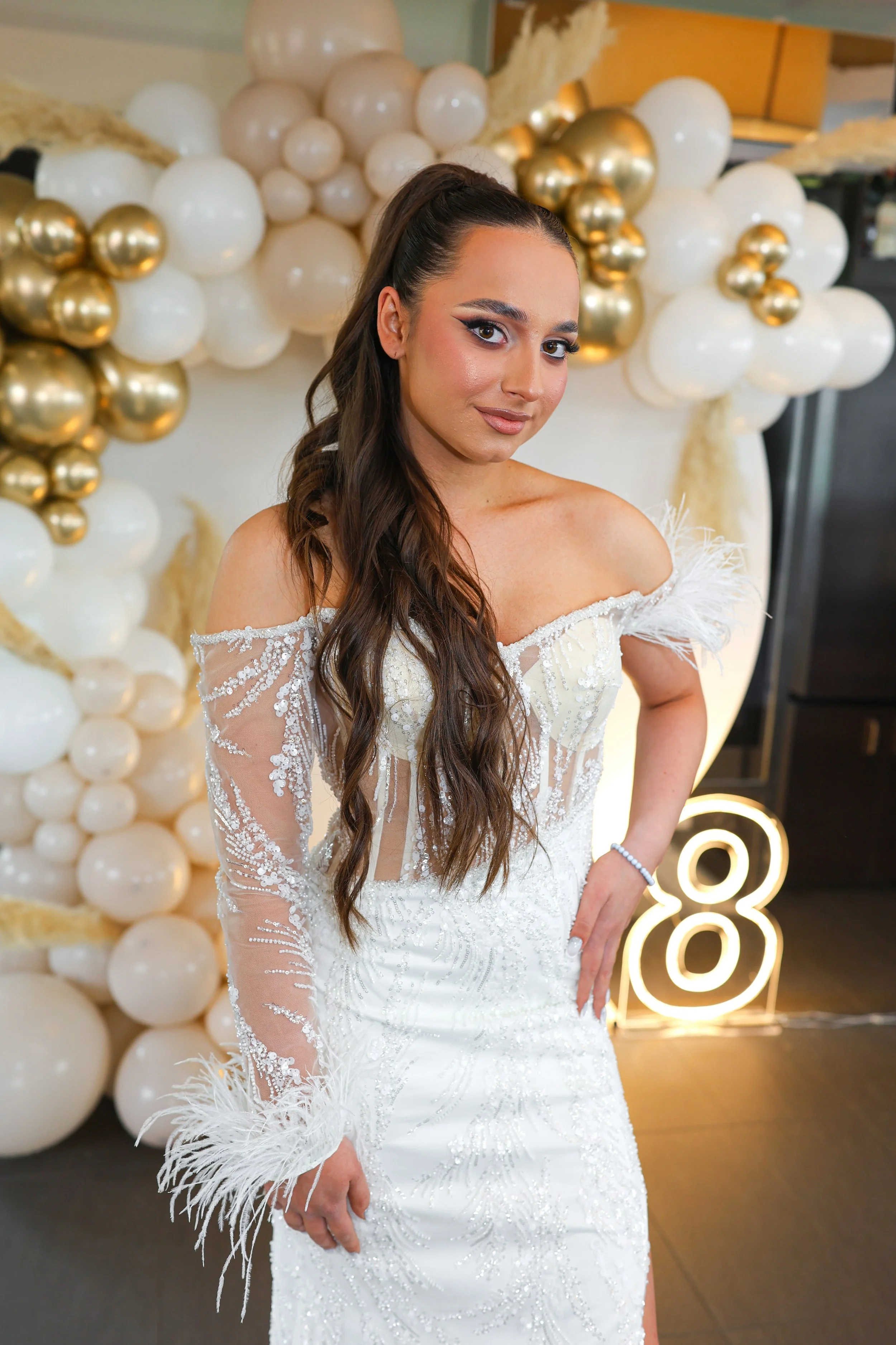 A woman in a white wedding dress with feather details and sheer, beaded sleeves, standing in front of a backdrop made of white and gold balloons.