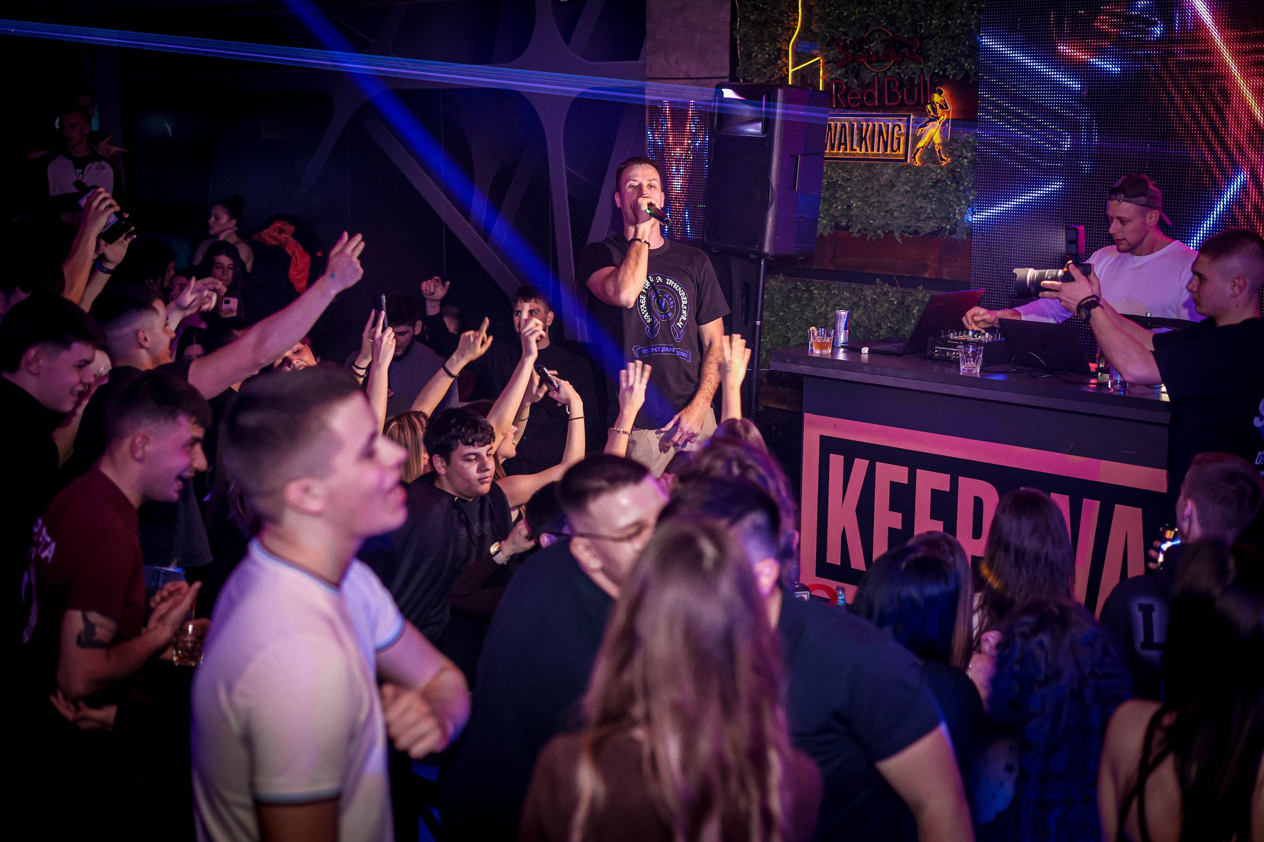 Crowd of young people at a nightclub or concert, facing a stage where a performer is speaking or singing into a microphone, with colorful lights and a DJ booth to the side.