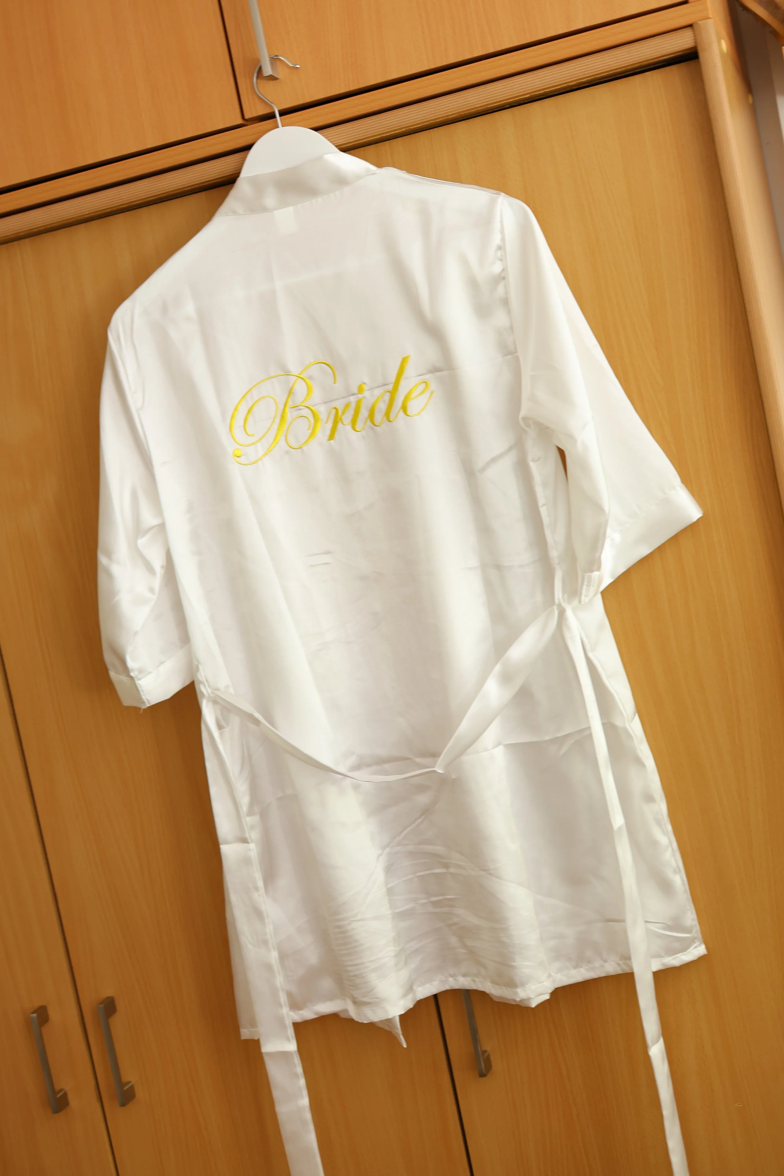 White satin robe hanging on a hanger on a wooden closet door with yellow embroidery that says 'Bride'.