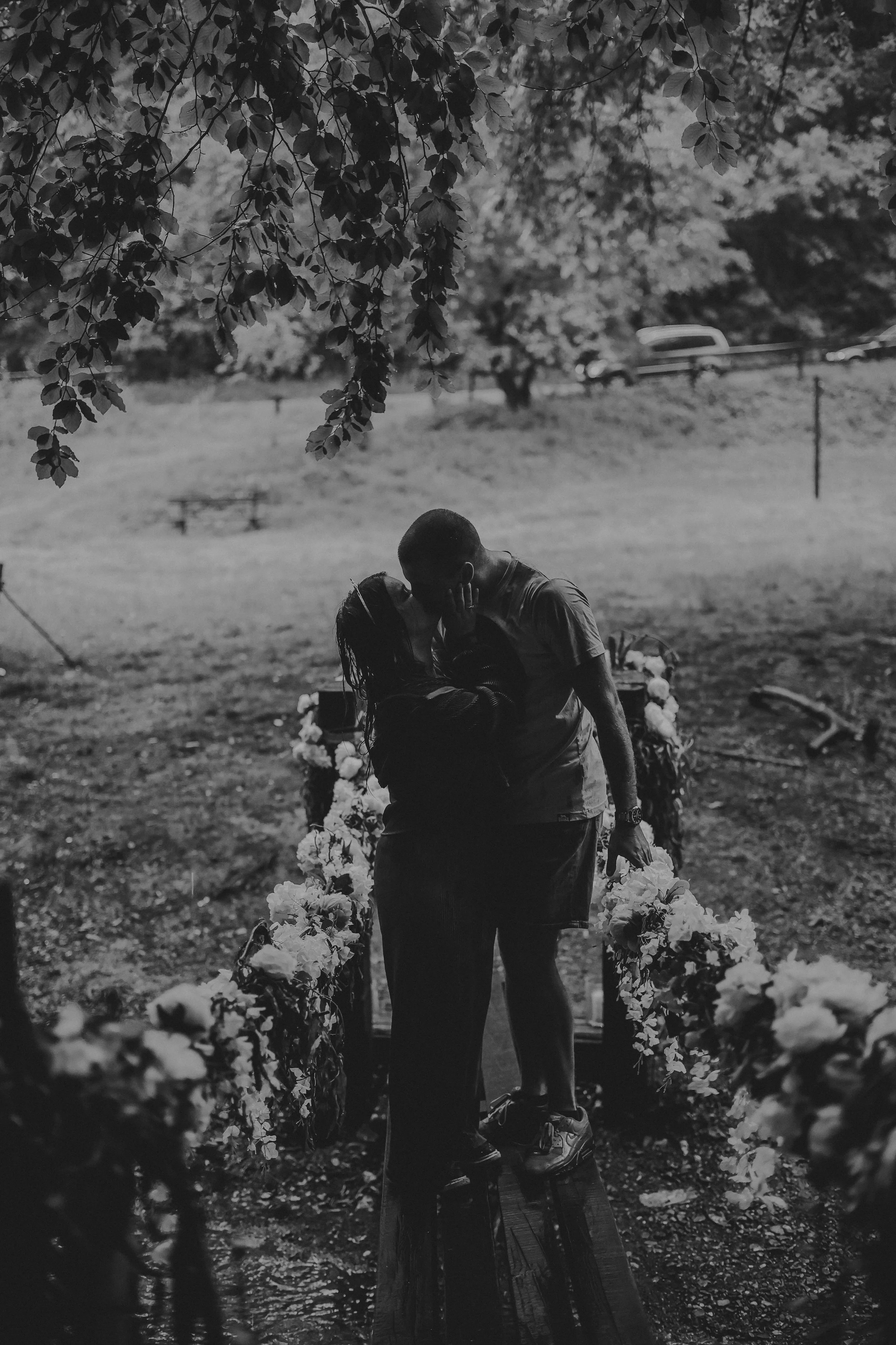A black and white photo of a couple kissing in a park, surrounded by flowers and trees.