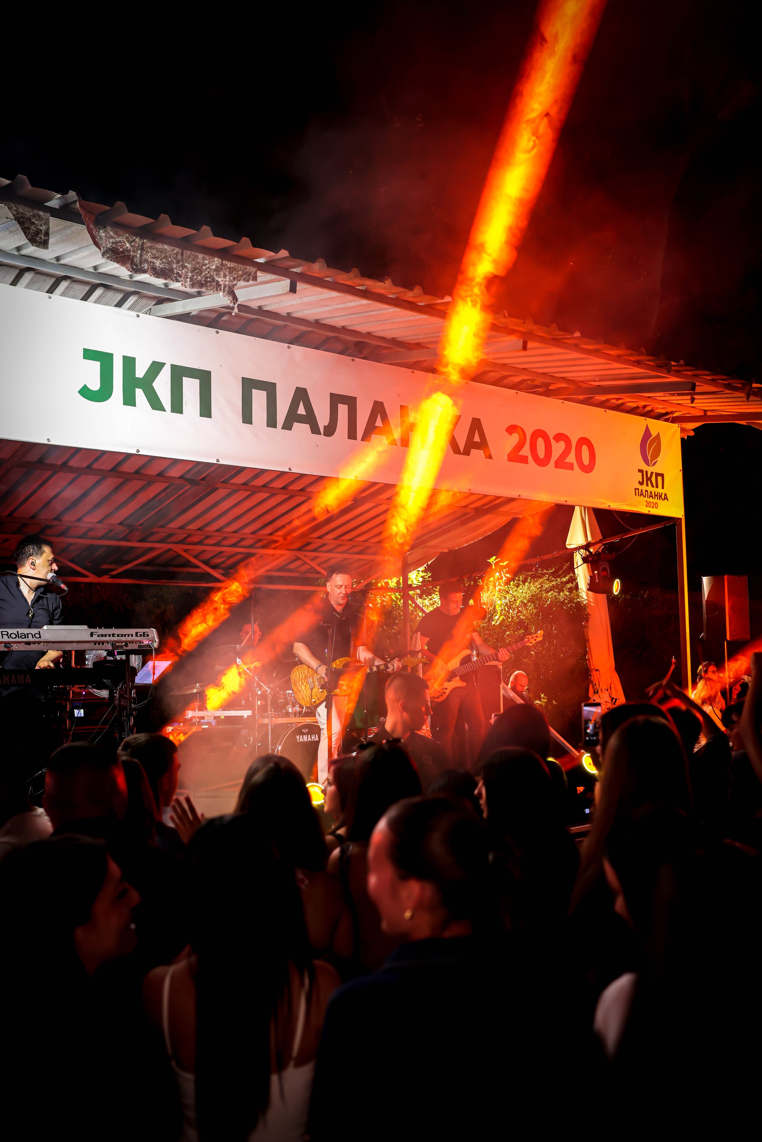 A band performs on stage at a night event, with bright orange flames shooting upward in the background and a crowd watching in the foreground. The stage has a sign with Greek text and the year 2020.
