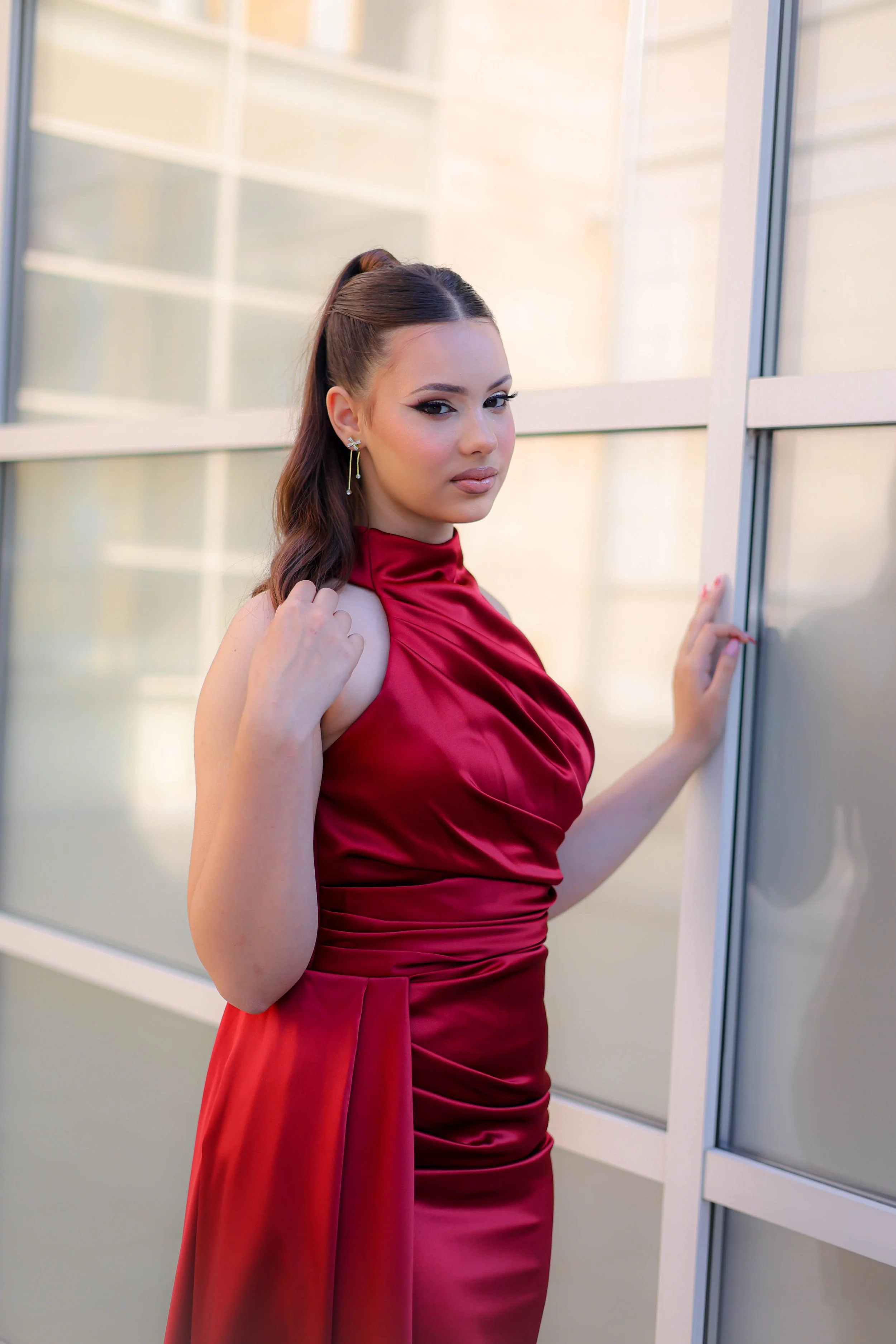 A woman with long brown hair tied in a high ponytail wearing a red satin dress, earrings, and makeup, standing by a glass window.