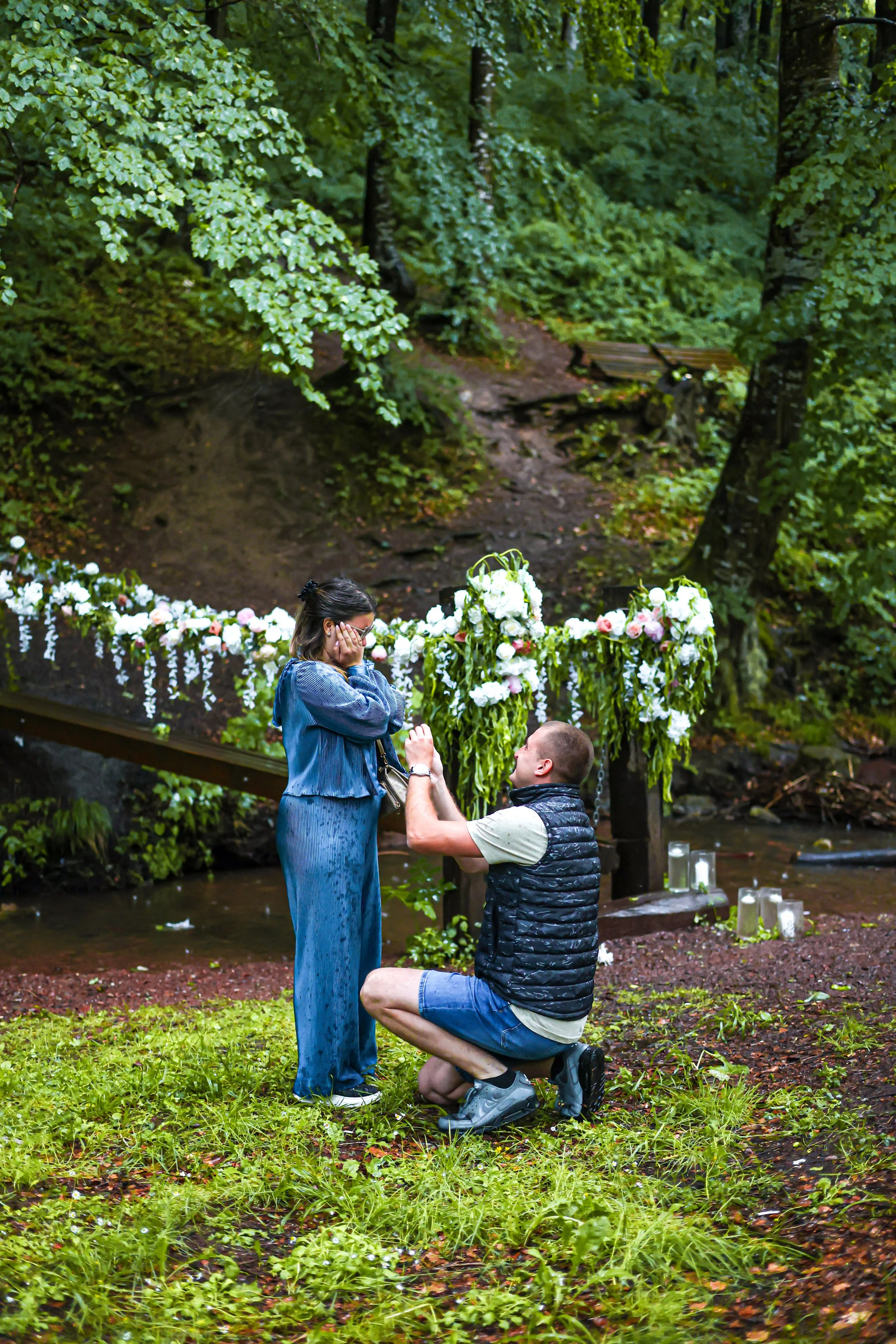 A man is proposing marriage to a woman in a forested area by a small stream, with flowers and candles decorate the scene.