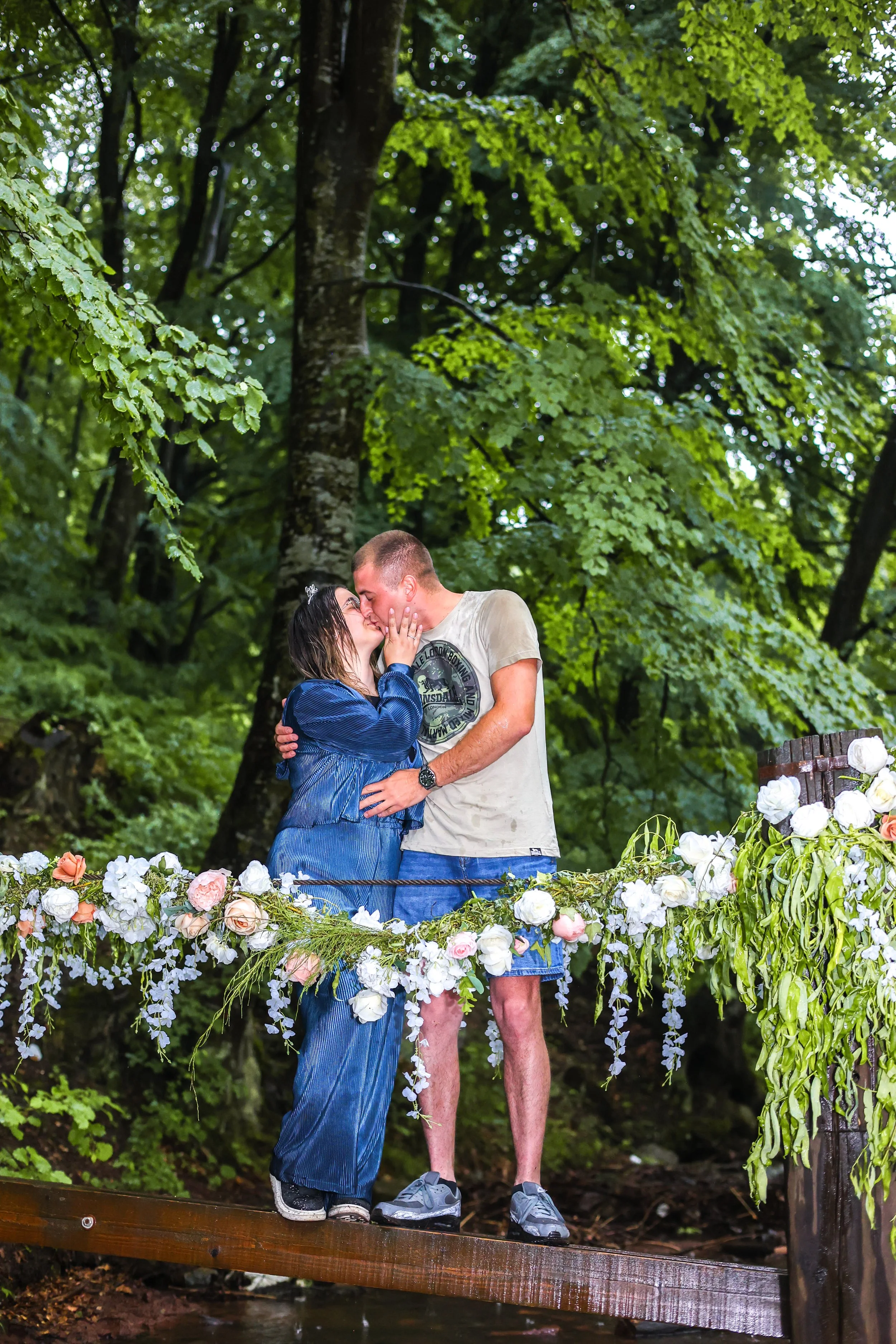 A couple kissing on a small wooden bridge decorated with white and pink flowers, surrounded by green trees.