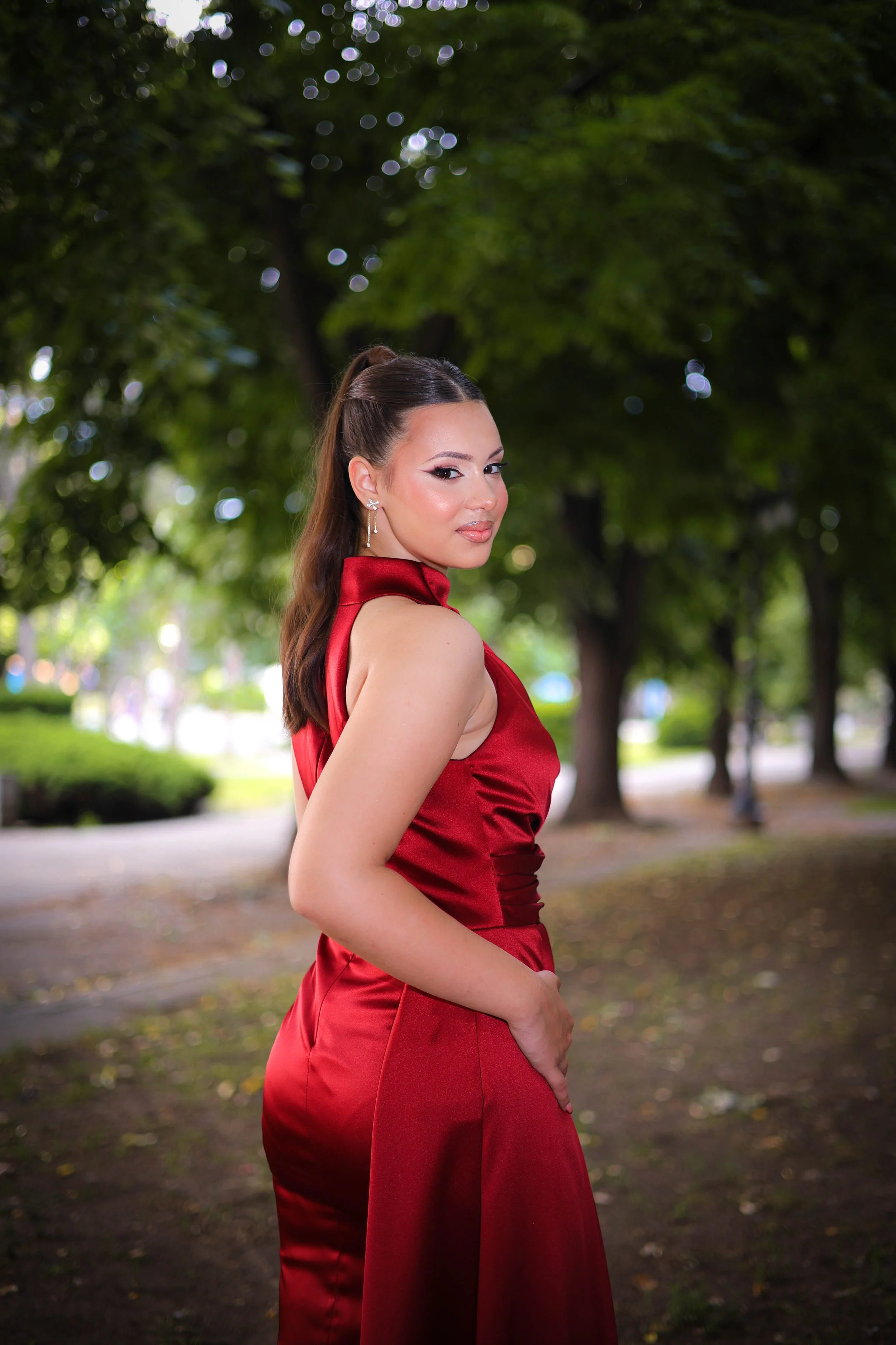 A woman in a red satin dress standing outdoors in a park with green trees in the background.