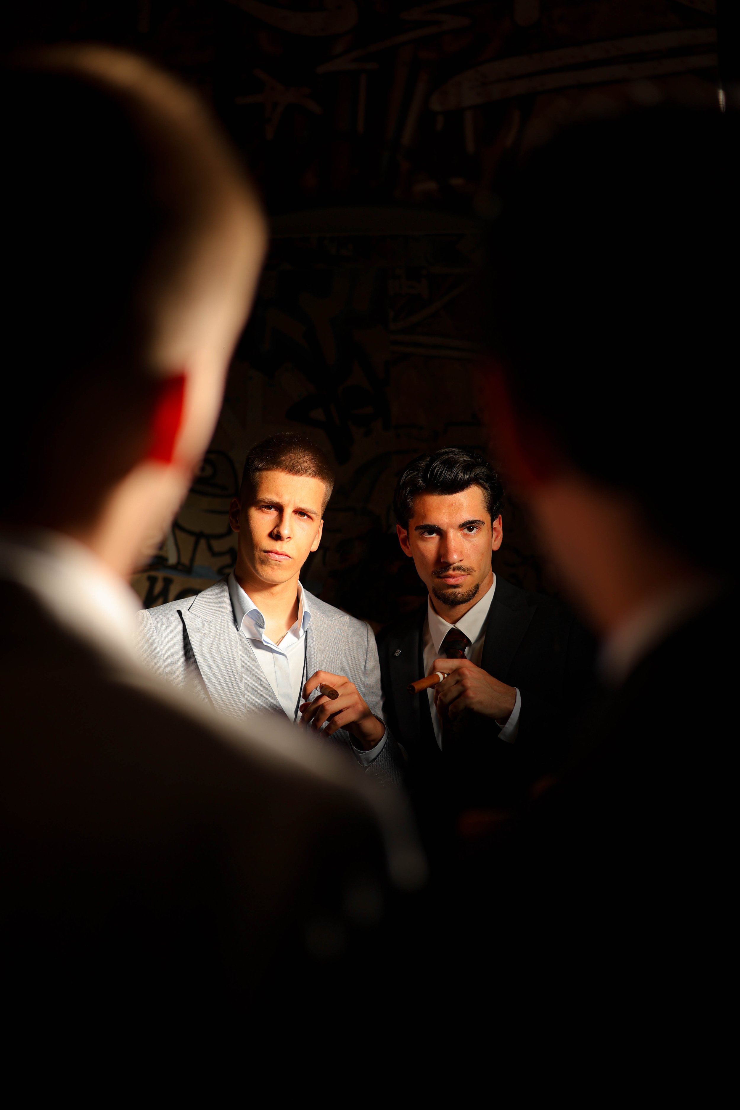 Two men in suits holding cigars, seen through an opening with blurred edges, with a graffiti-covered wall in the background.
