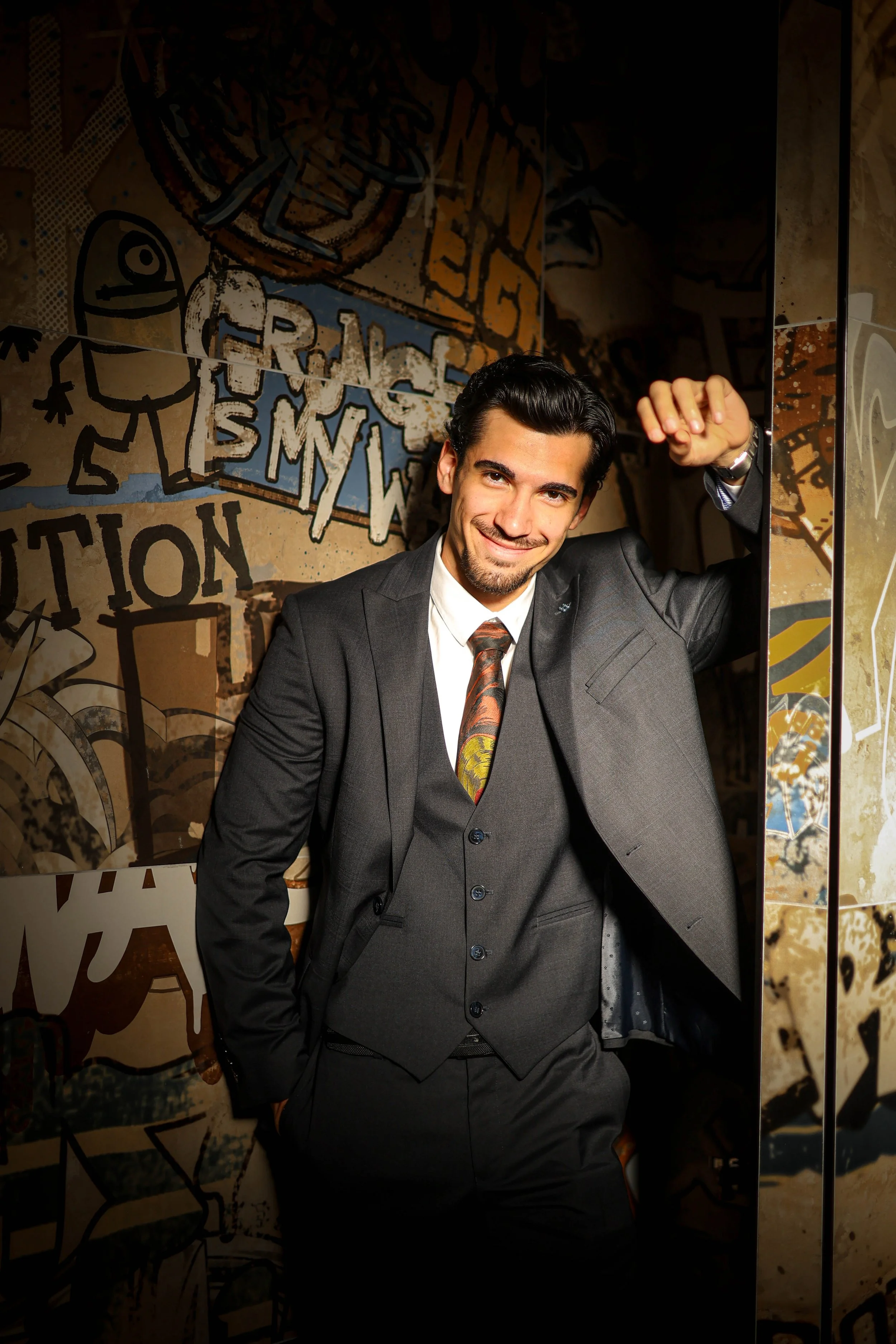 A young man in a dark suit with a white shirt and patterned tie, smiling and leaning against a wall with graffiti art and text behind him.