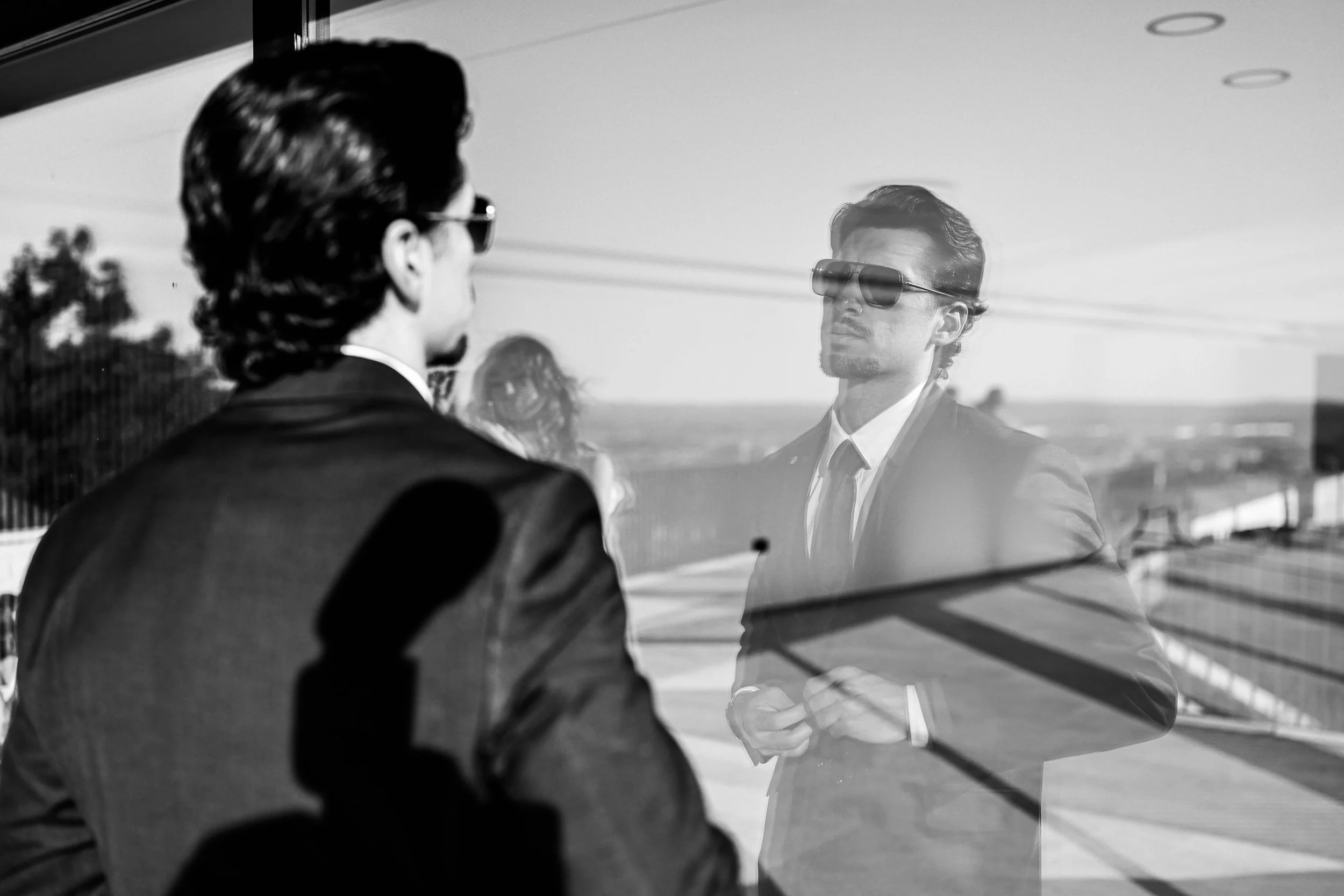 A man with sunglasses and styled hair is seen through a glass window, with his reflection visible on the surface. The scene is in black and white.