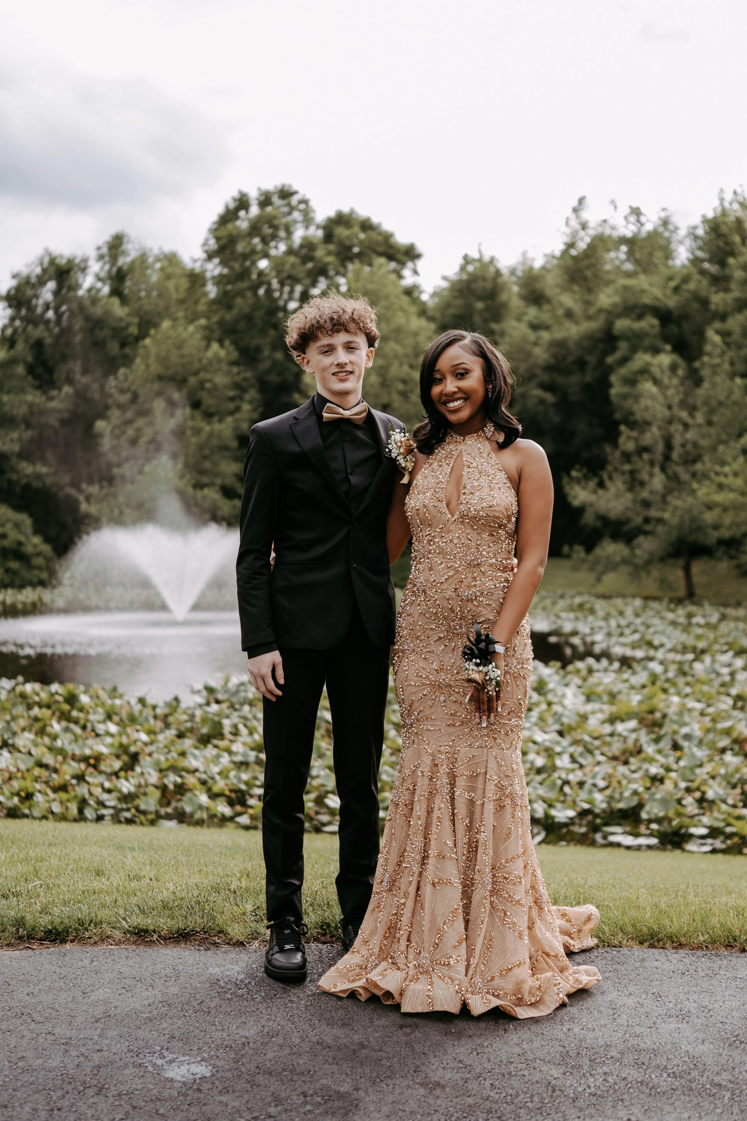 A young couple dressed in formal attire standing outdoors in front of a pond with a fountain and lush greenery, smiling for the camera.