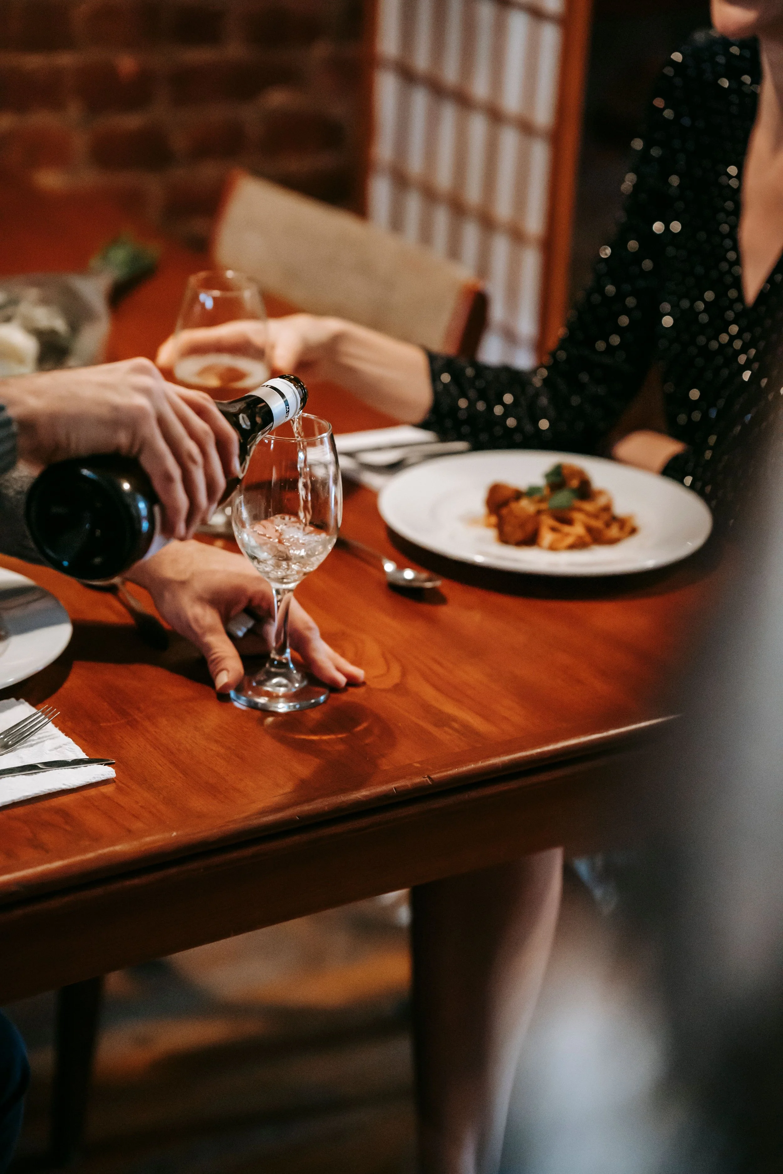 People having a meal at a wooden table, with one person pouring a drink into a glass and another person sitting with a plate of pasta.