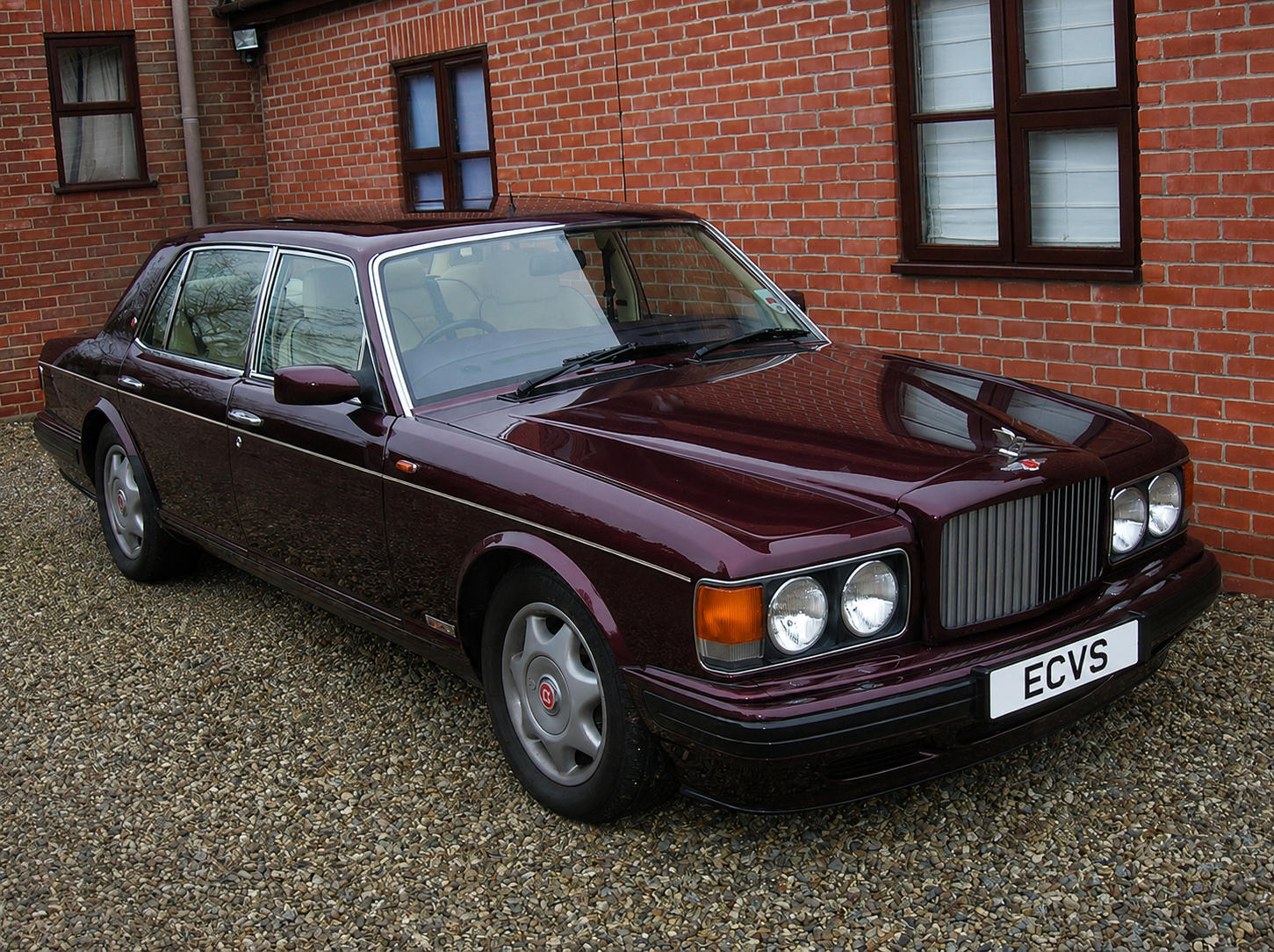 A vintage dark red luxury sedan car parked on gravel in front of a brick house with brown-framed windows.