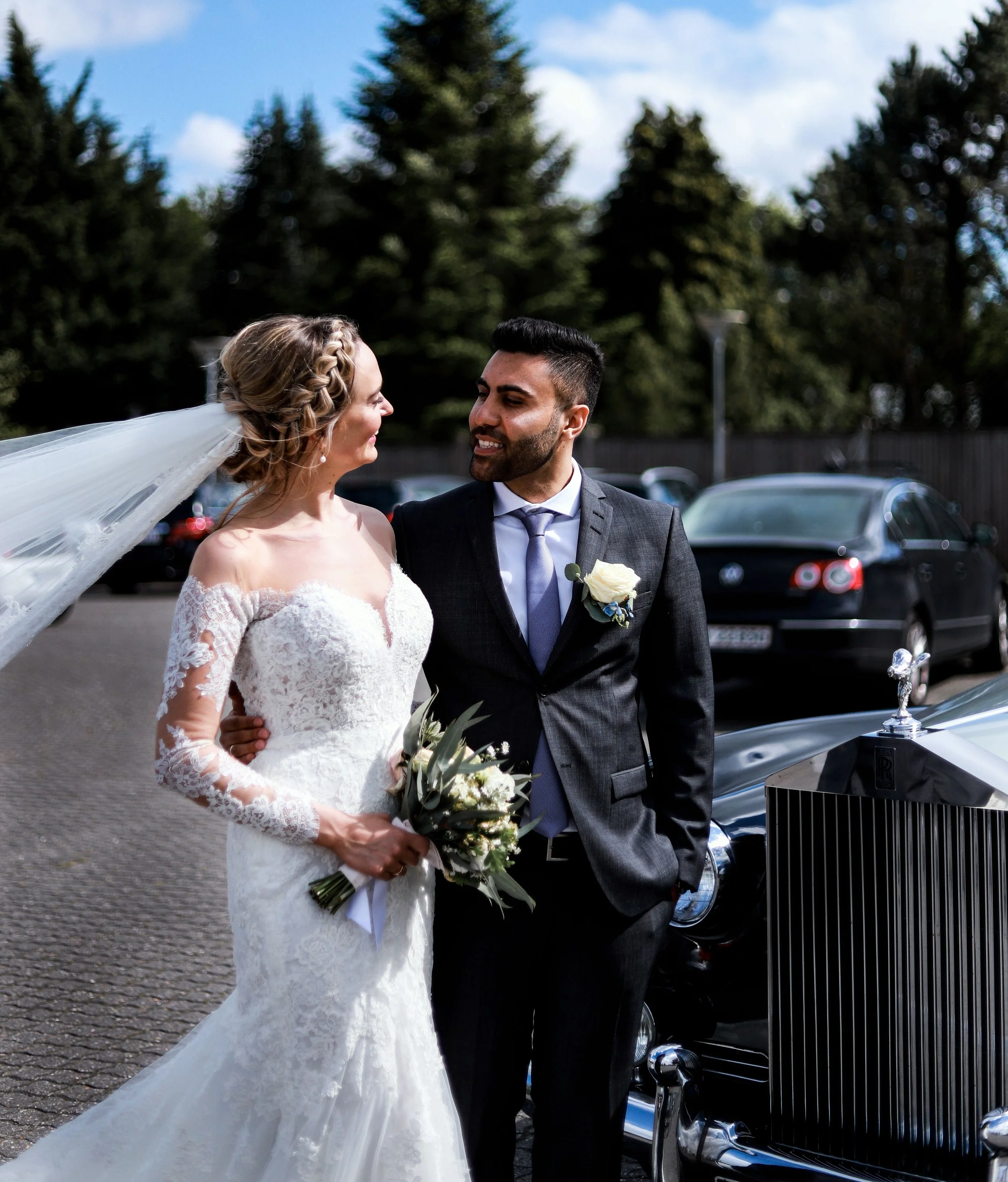 A bride and groom standing close to each other outdoors, smiling and gazing into each other's eyes, with the bride holding a bouquet of flowers, dressed in a lace wedding gown and veil, against a background of trees and parked cars.