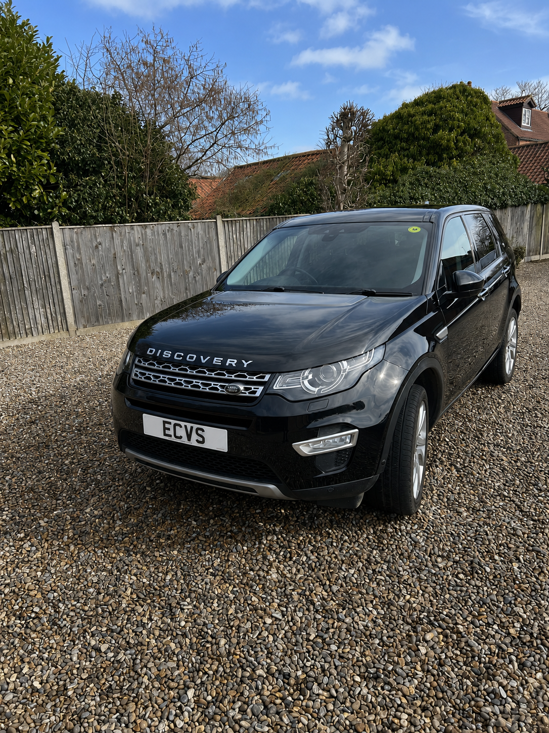 A black Land Rover Discovery SUV parked on a gravel driveway with a wooden fence, trees, and houses in the background.