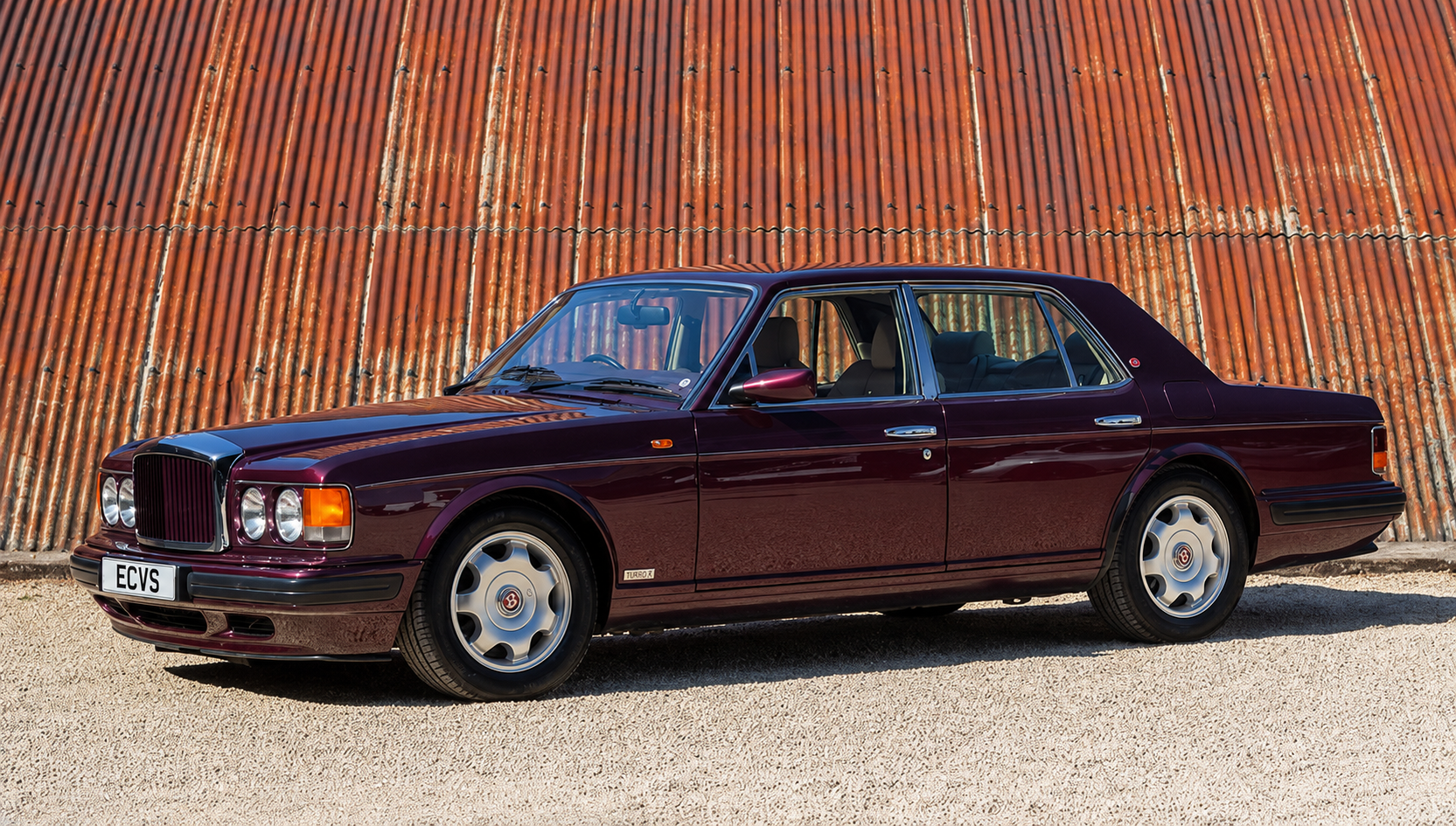 A classic maroon luxury sedan parked in front of a weathered, rust-colored corrugated metal wall. The car features a boxy design with round headlights, silver hubcaps, and a sleek body.