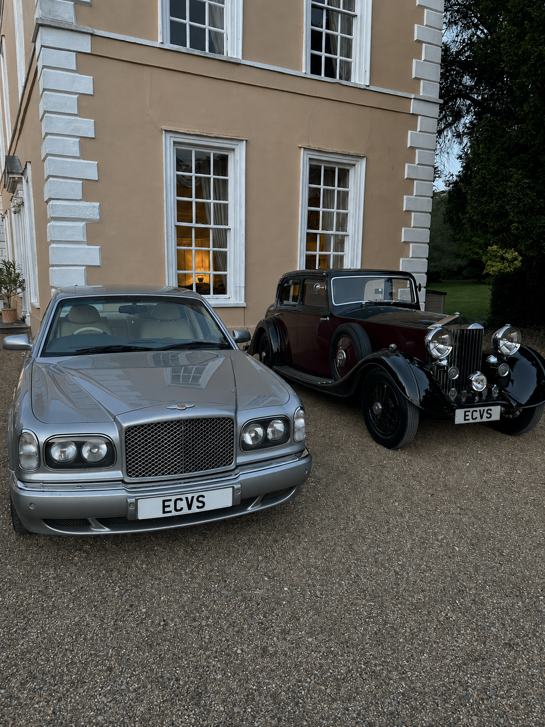 A silver modern luxury car parked beside a vintage black and burgundy automobile in front of a yellow building with white window frames.