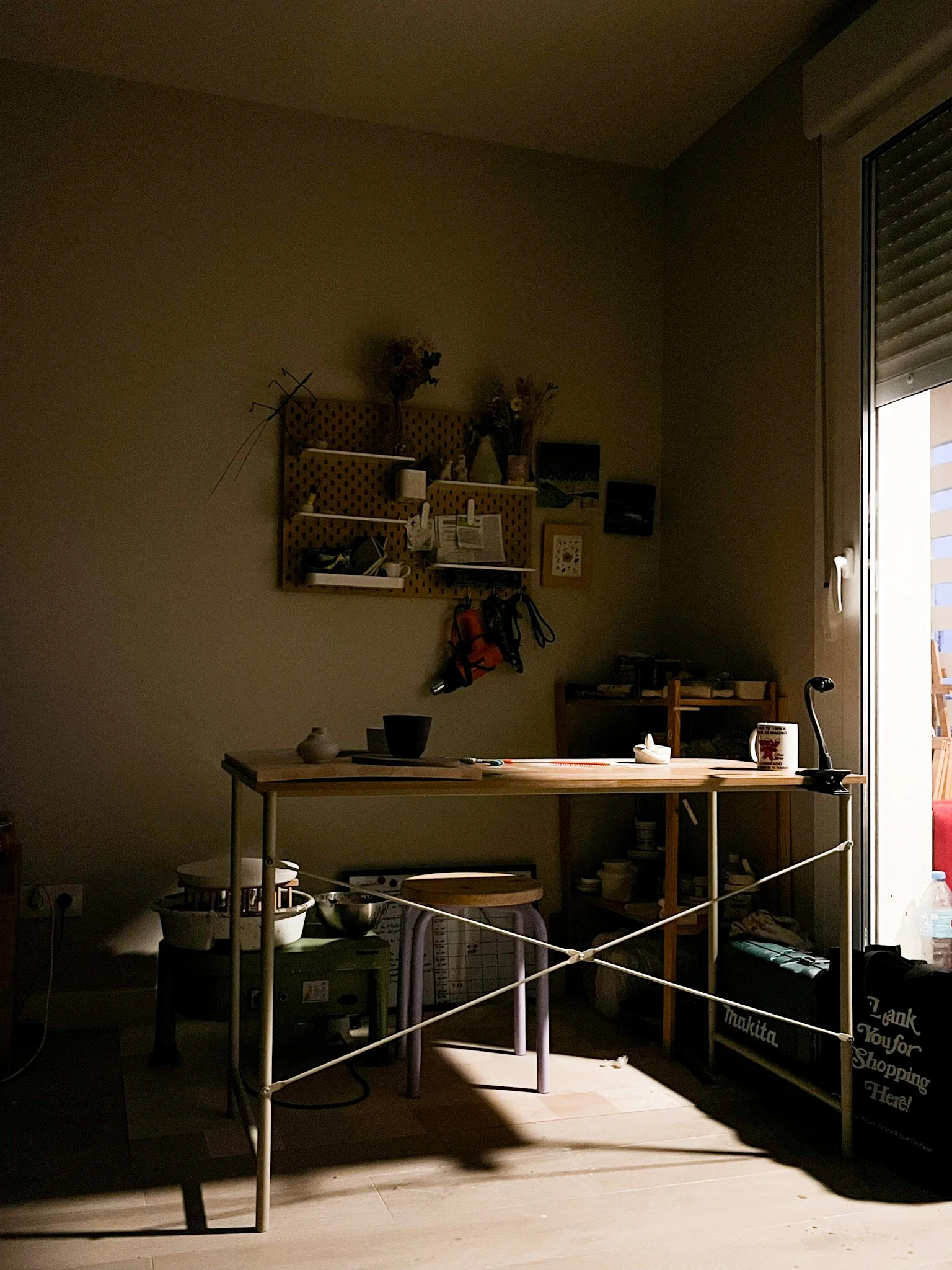 Interior de una habitación con una mesa, estantería y objetos dispersos, al lado de una ventana con cortinas Horizontal y luz natural entrando.