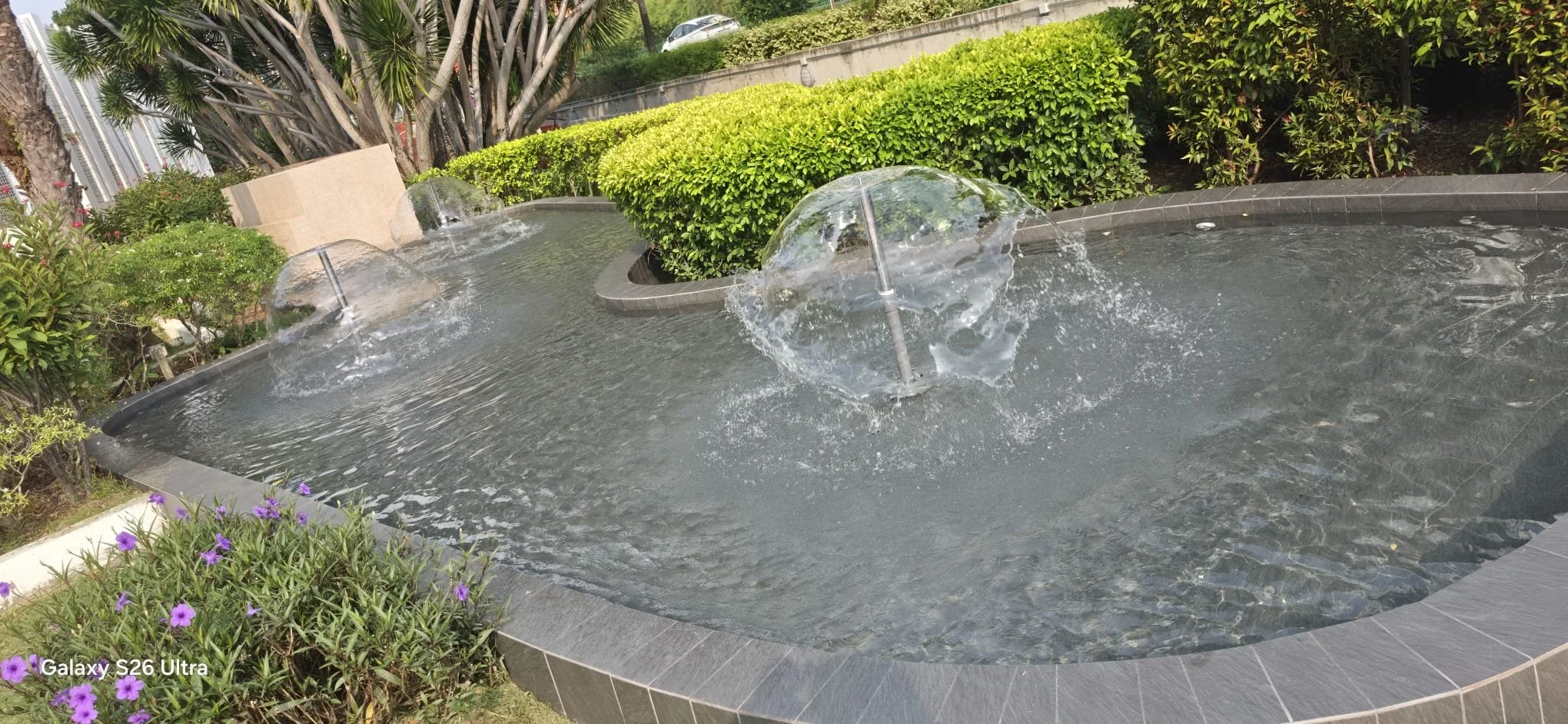 A rectangular fountain with three water jets shooting water into the air, surrounded by green bushes, purple flowers, and trees in an outdoor setting.